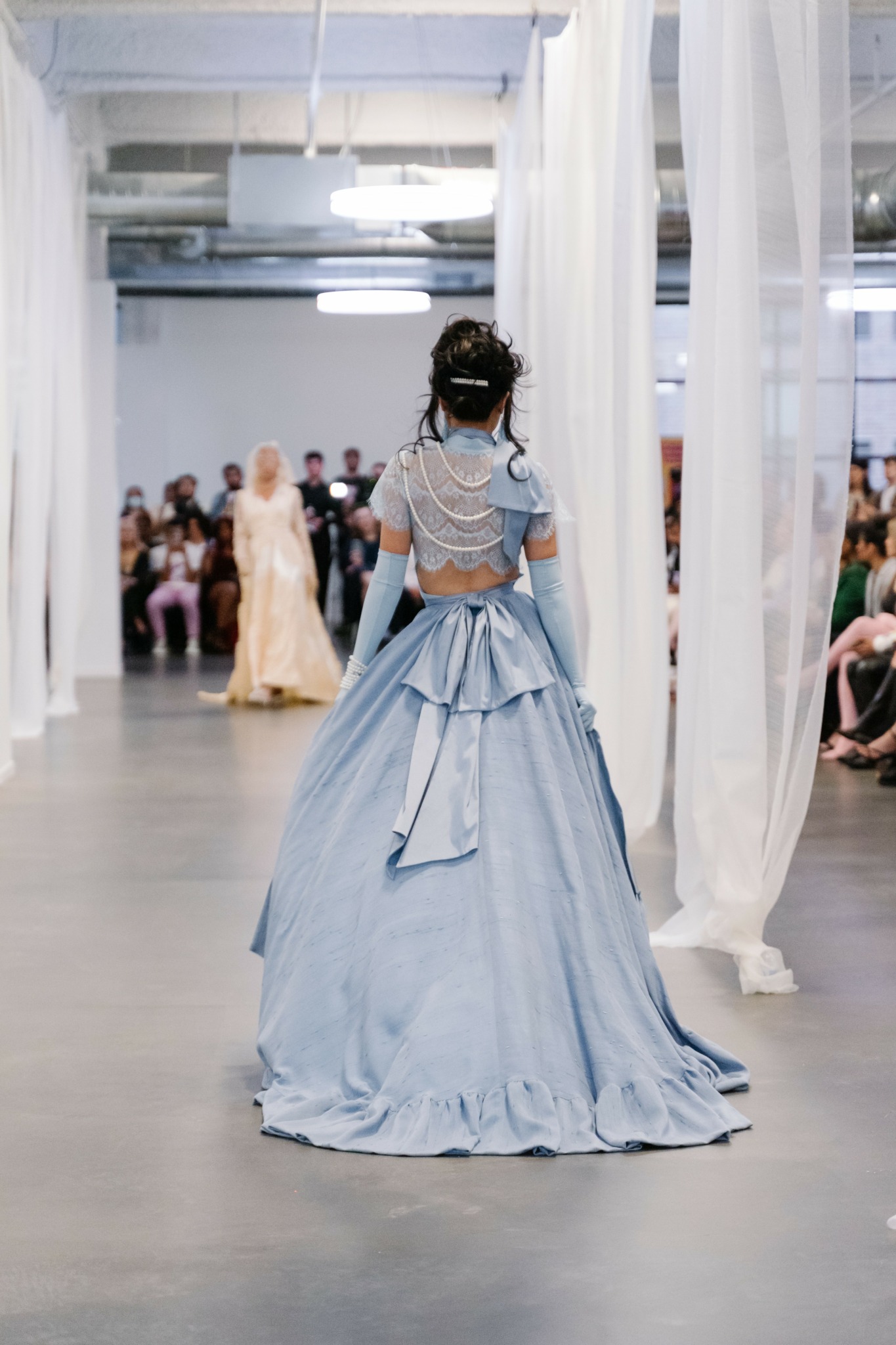 Person in a blue gown walking down a runway with audience on both sides, white curtains hanging around.