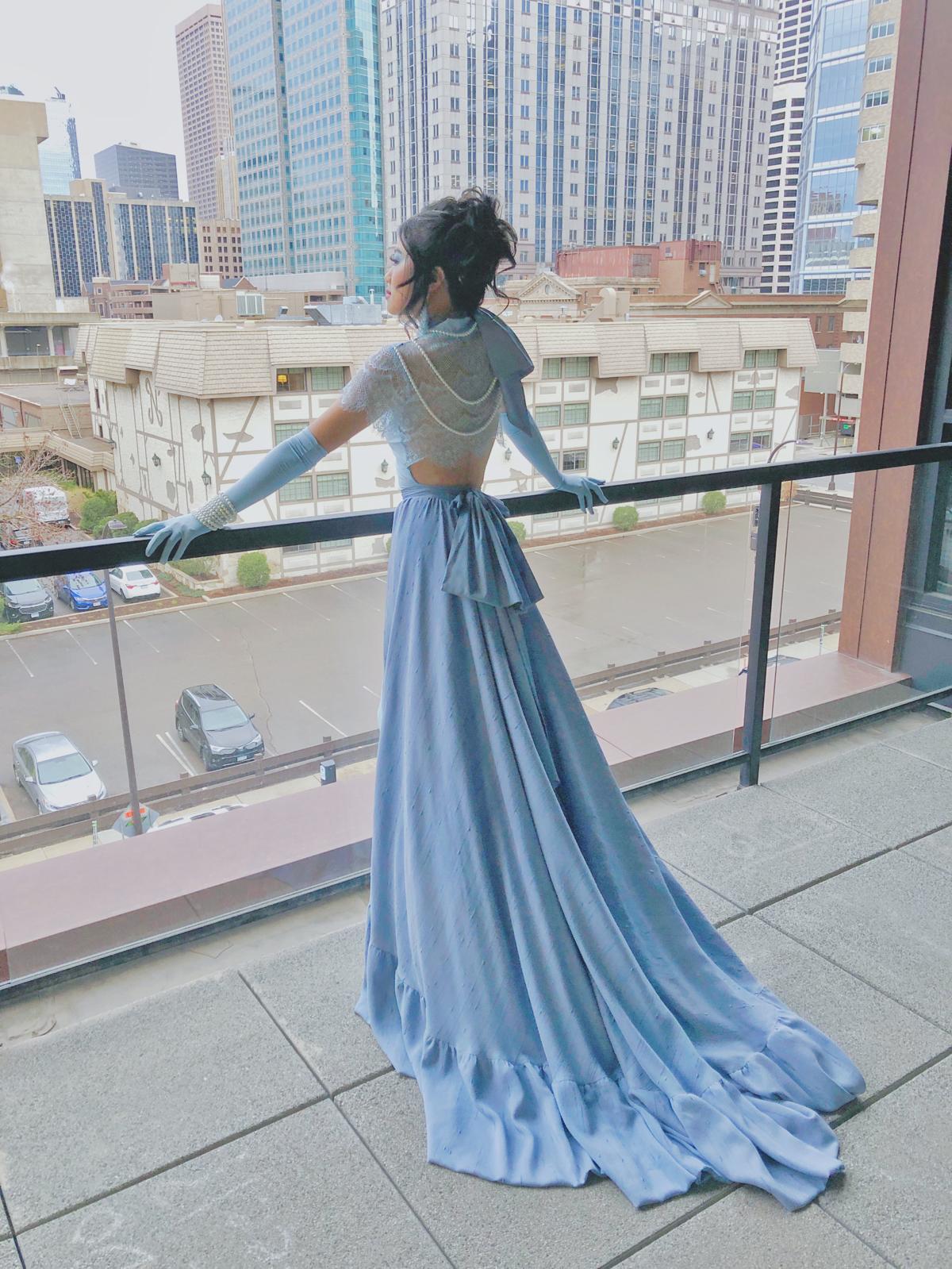 Woman in a long, flowing blue dress standing on a balcony overlooking city buildings and streets.