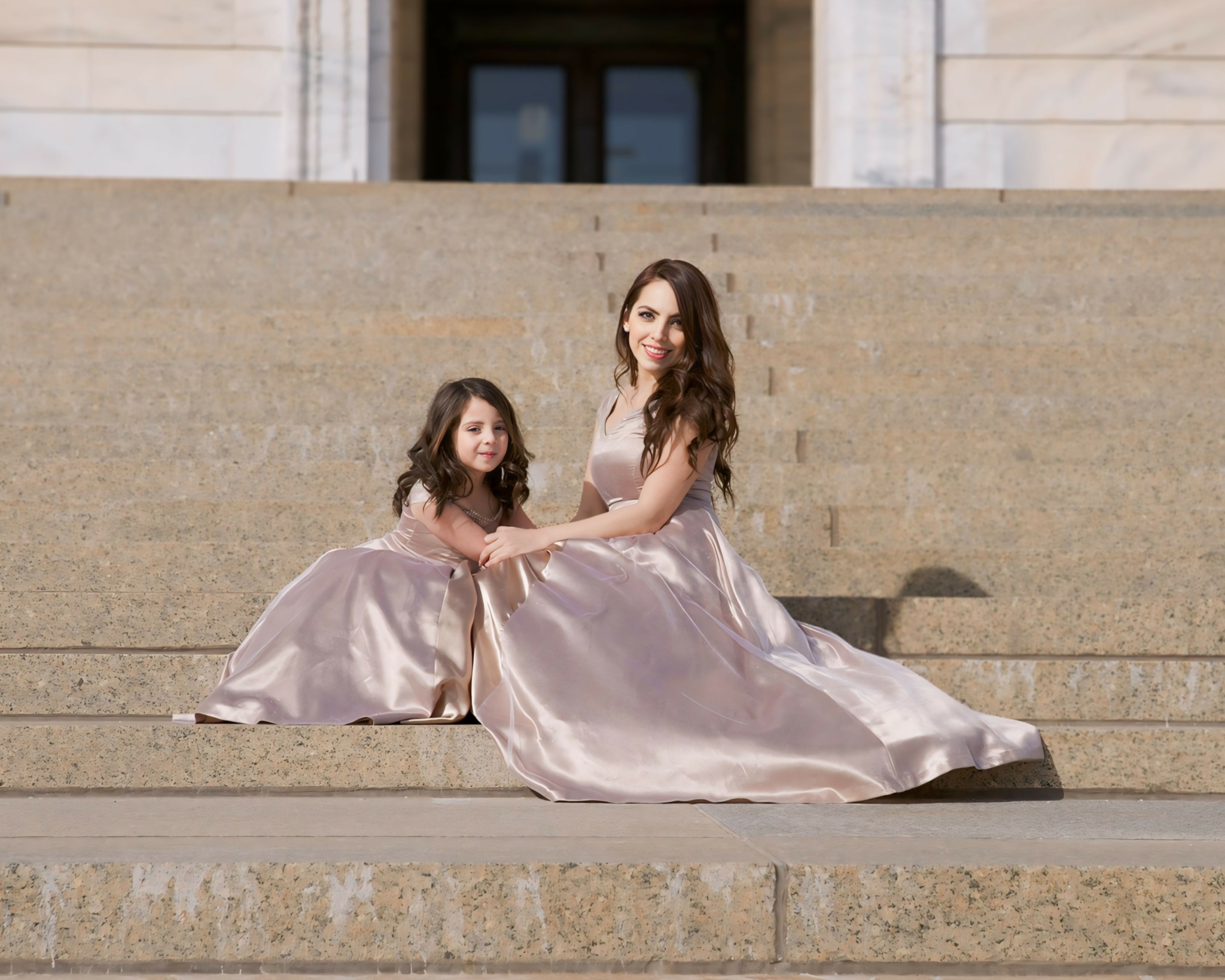 Two females in matching satin dresses sitting on stone steps outside building, smiling at camera.