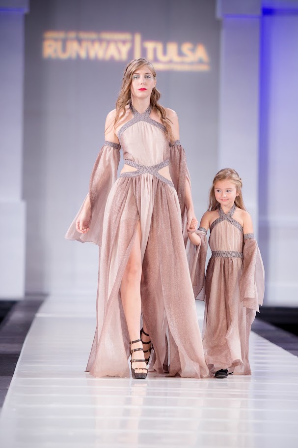 Two models walk down runway in flowing, pastel-colored dresses, holding hands, with runway and backdrop in background.