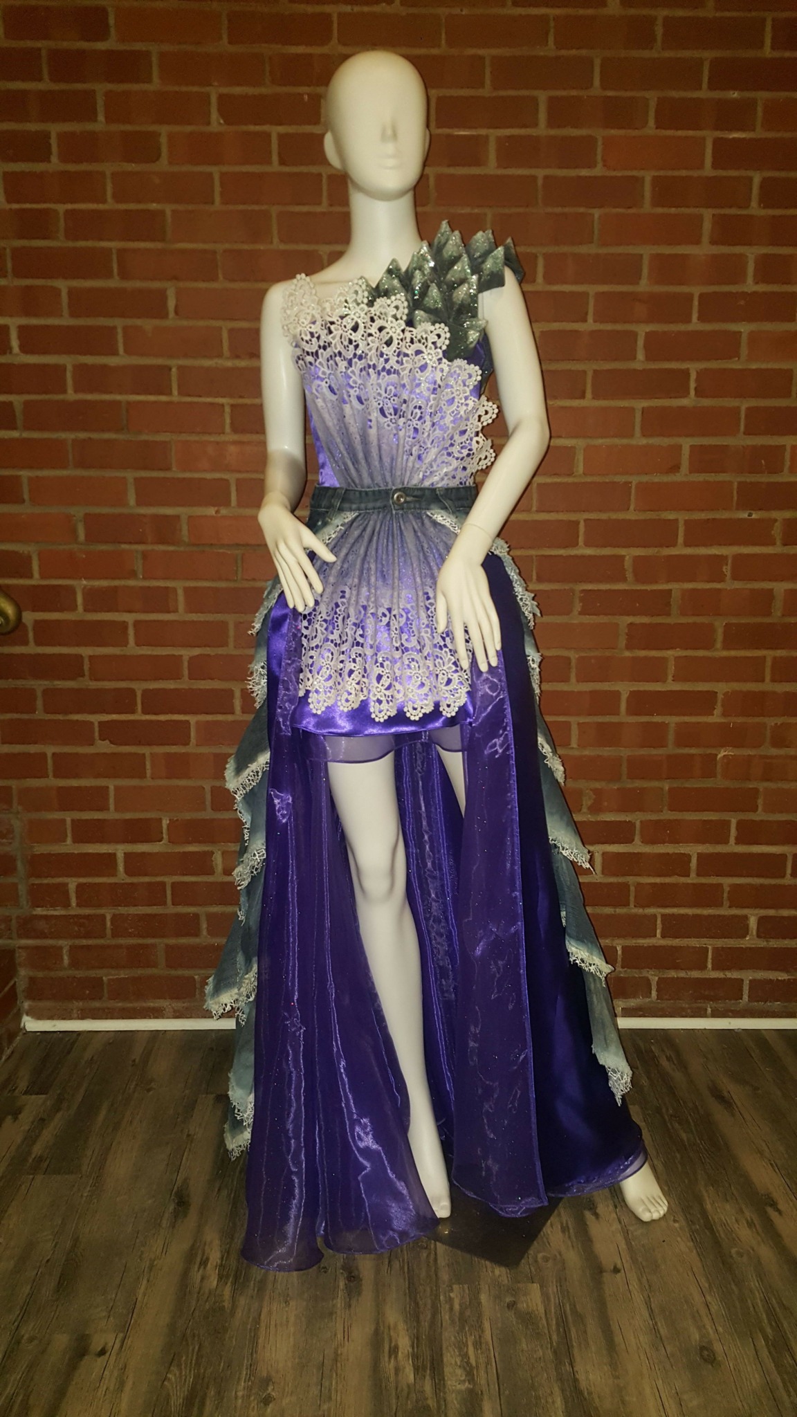 Mannequin wearing a purple and lavender gown with floral details, standing against a brick wall.