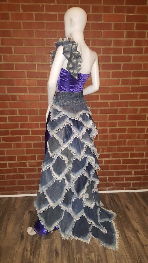 Mannequin wearing a purple and gray layered gown with ruffles, standing against a brick wall.
