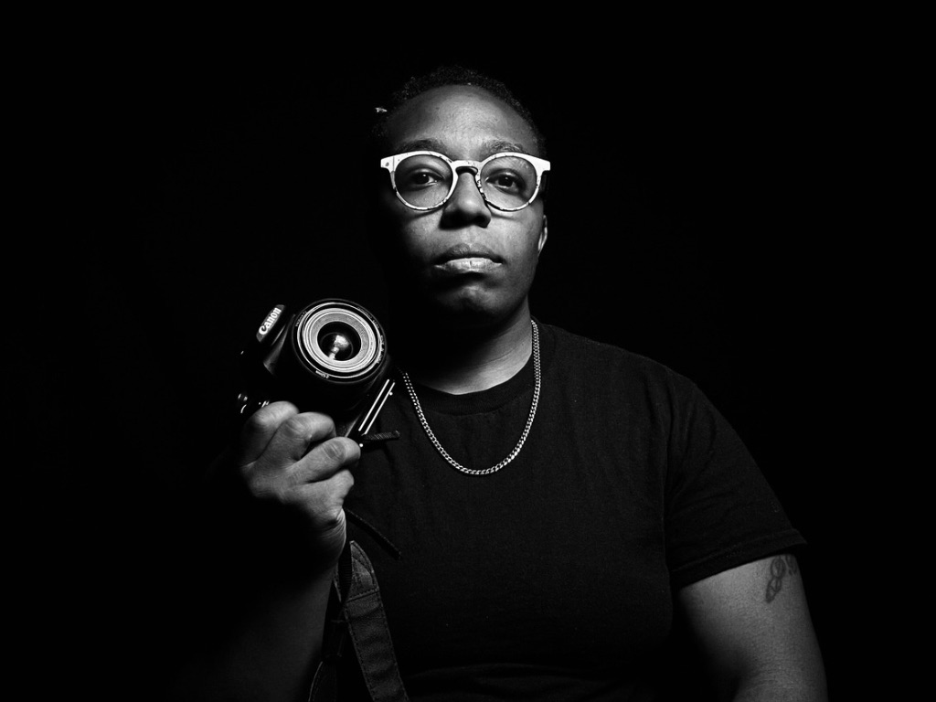 Person with glasses holding a camera, looking directly at the camera, dark background.