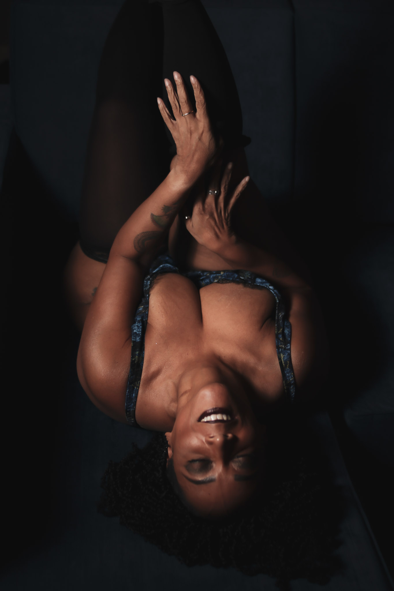 Woman lying on dark surface, smiling, wearing a blue top, with arms raised and hands near face.