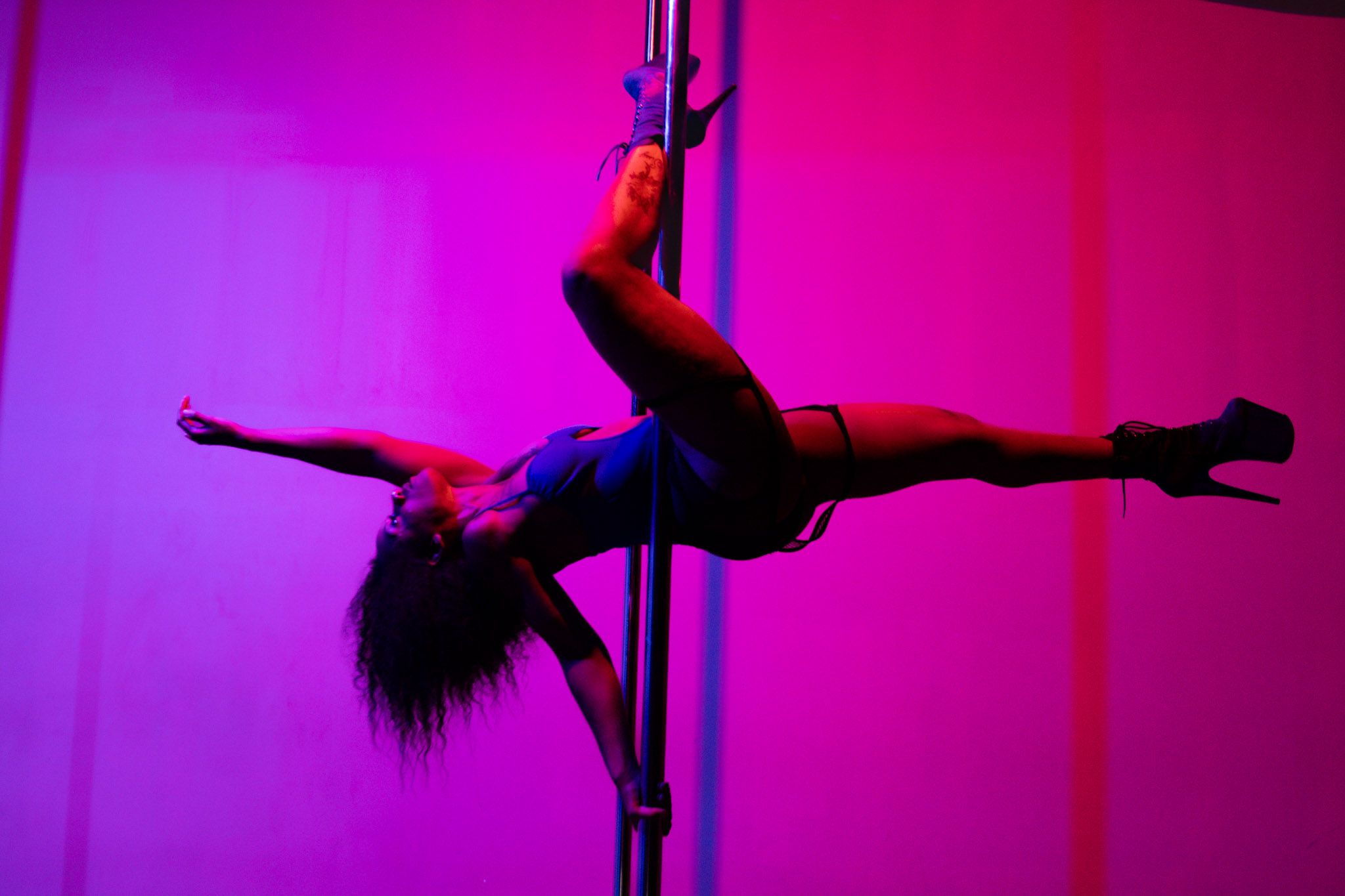 Performer hangs horizontally on a pole with arms and legs extended, colorful stage lighting in background.