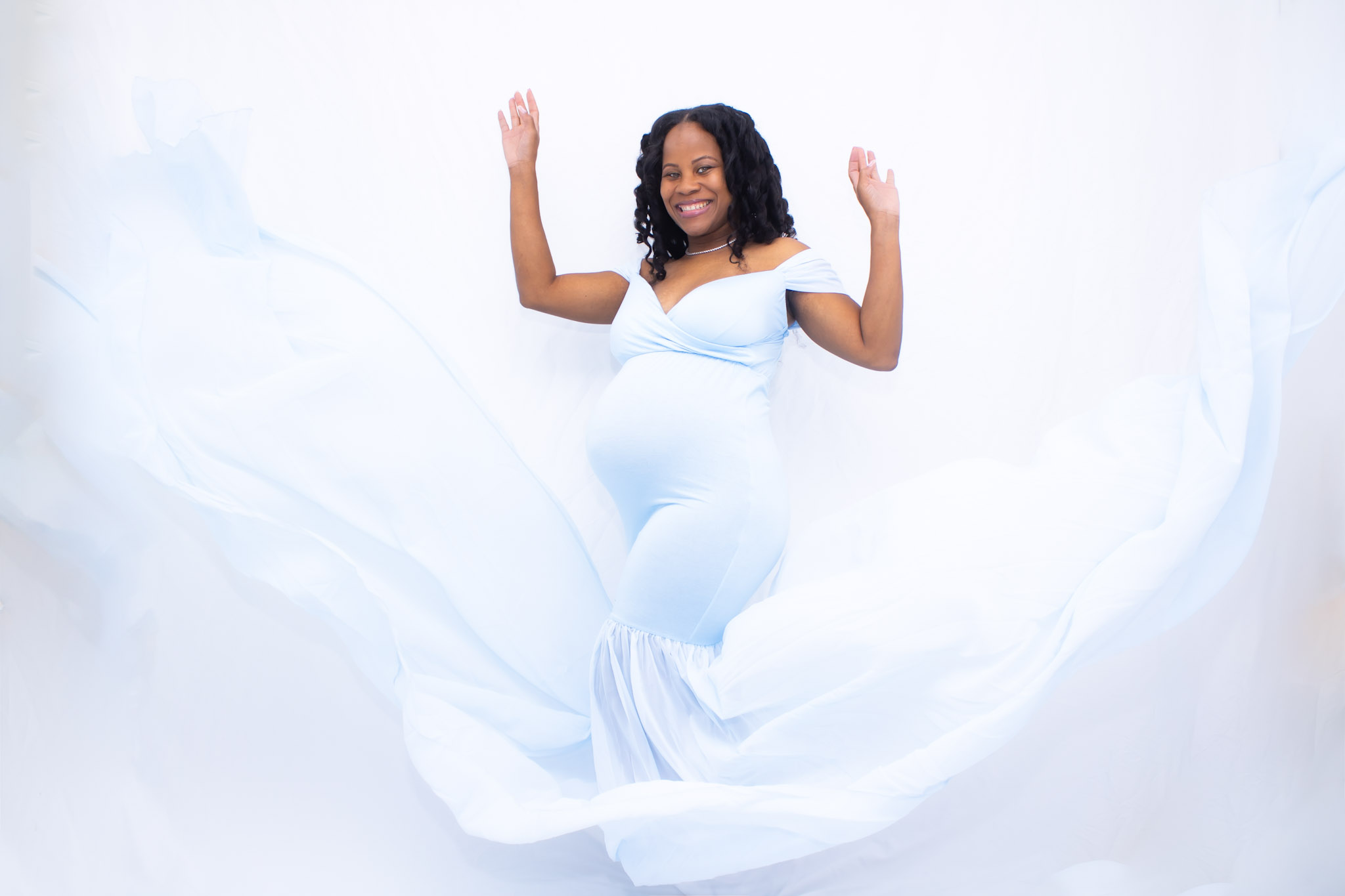 Pregnant woman in a white dress smiling with arms raised, surrounded by flowing white fabric, against a white background.