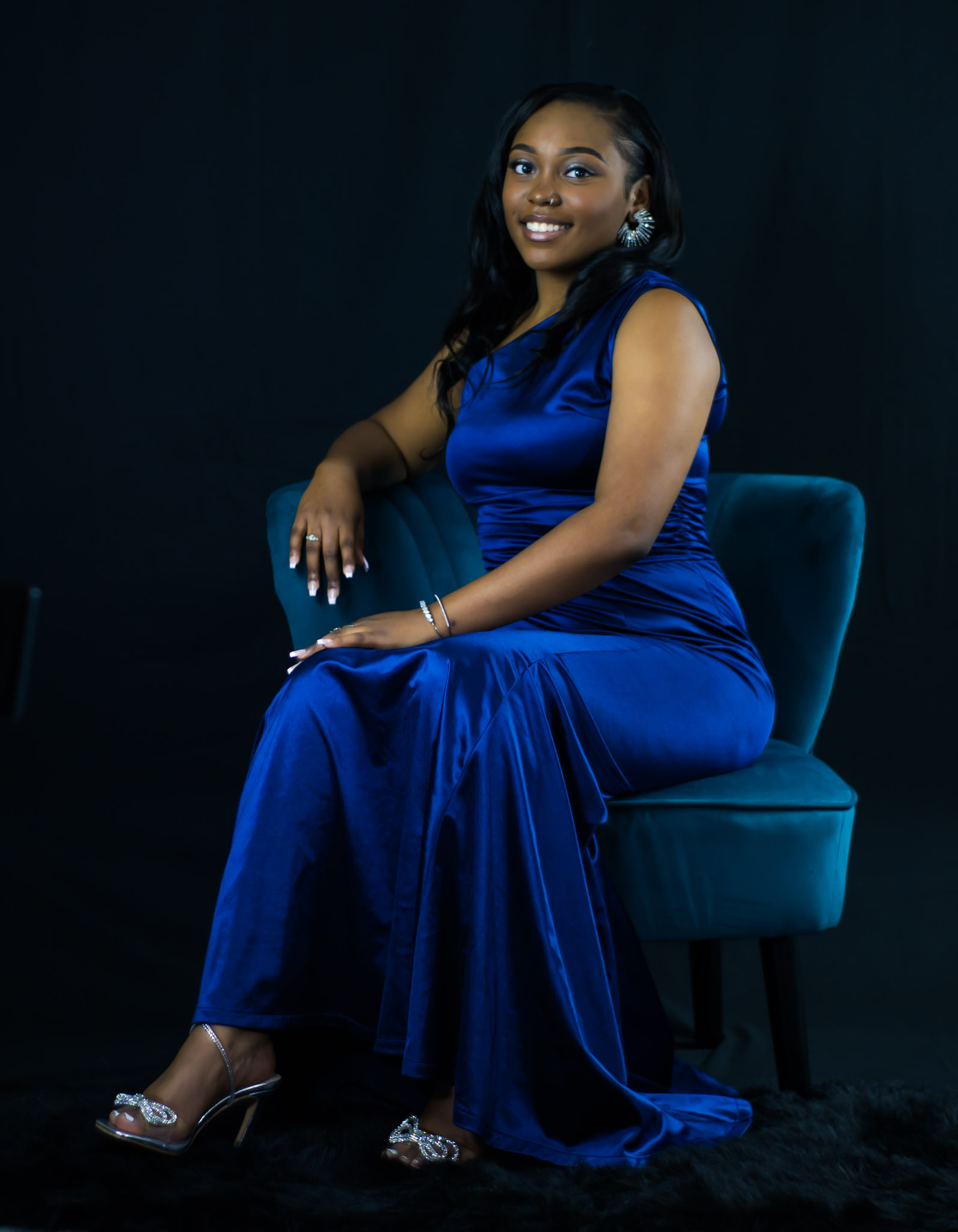 Woman in a blue dress sitting on a teal chair, smiling, dark background, elegant pose.