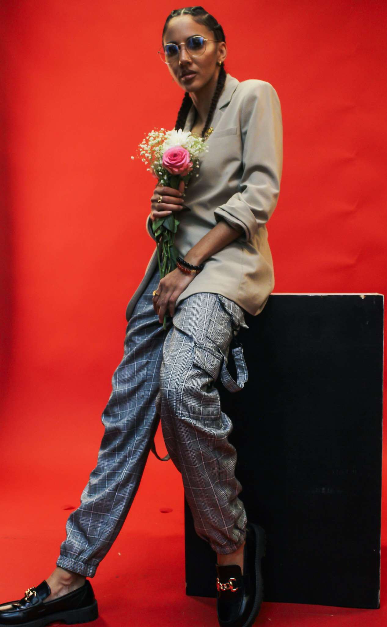 Person holding a bouquet of flowers, standing against a red background, wearing glasses, a beige jacket, and patterned pants.