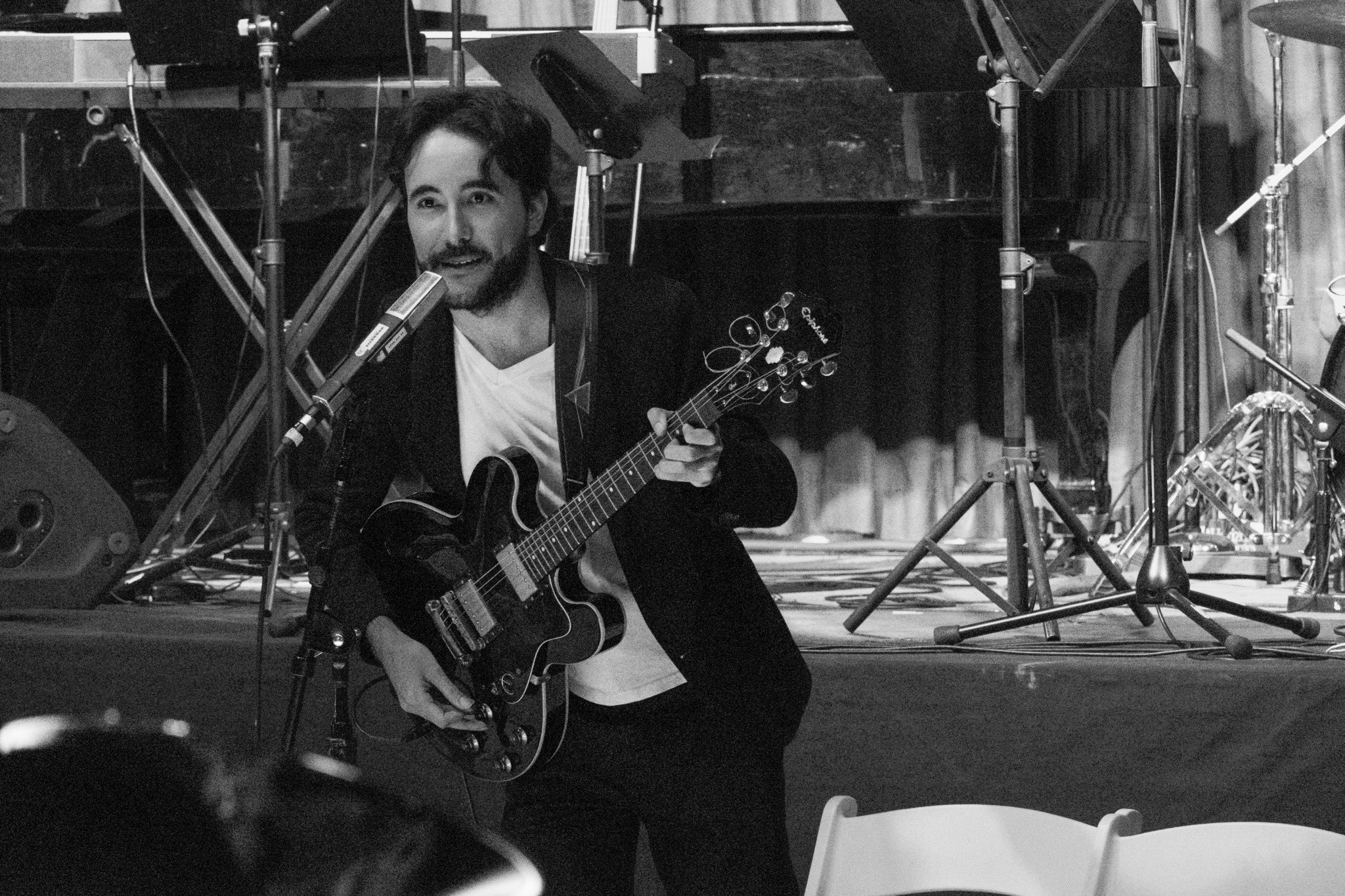 Musician with guitar performing on stage with microphone and musical equipment in background.