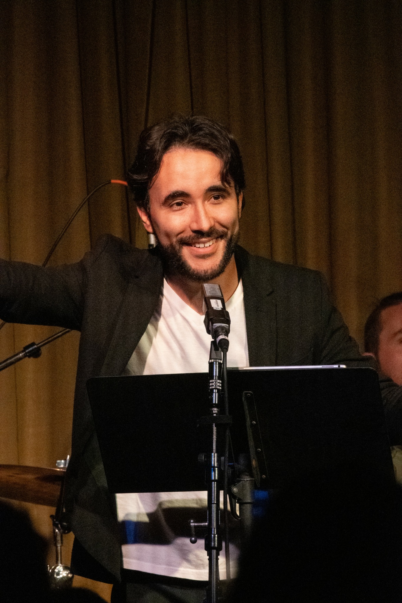 Man with dark hair and beard smiling, standing behind a microphone, in front of a dark curtain.