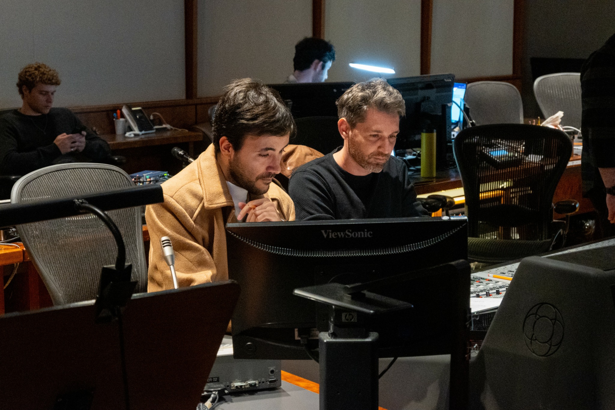 Two men sit at a desk working on a computer in a room with multiple workstations.