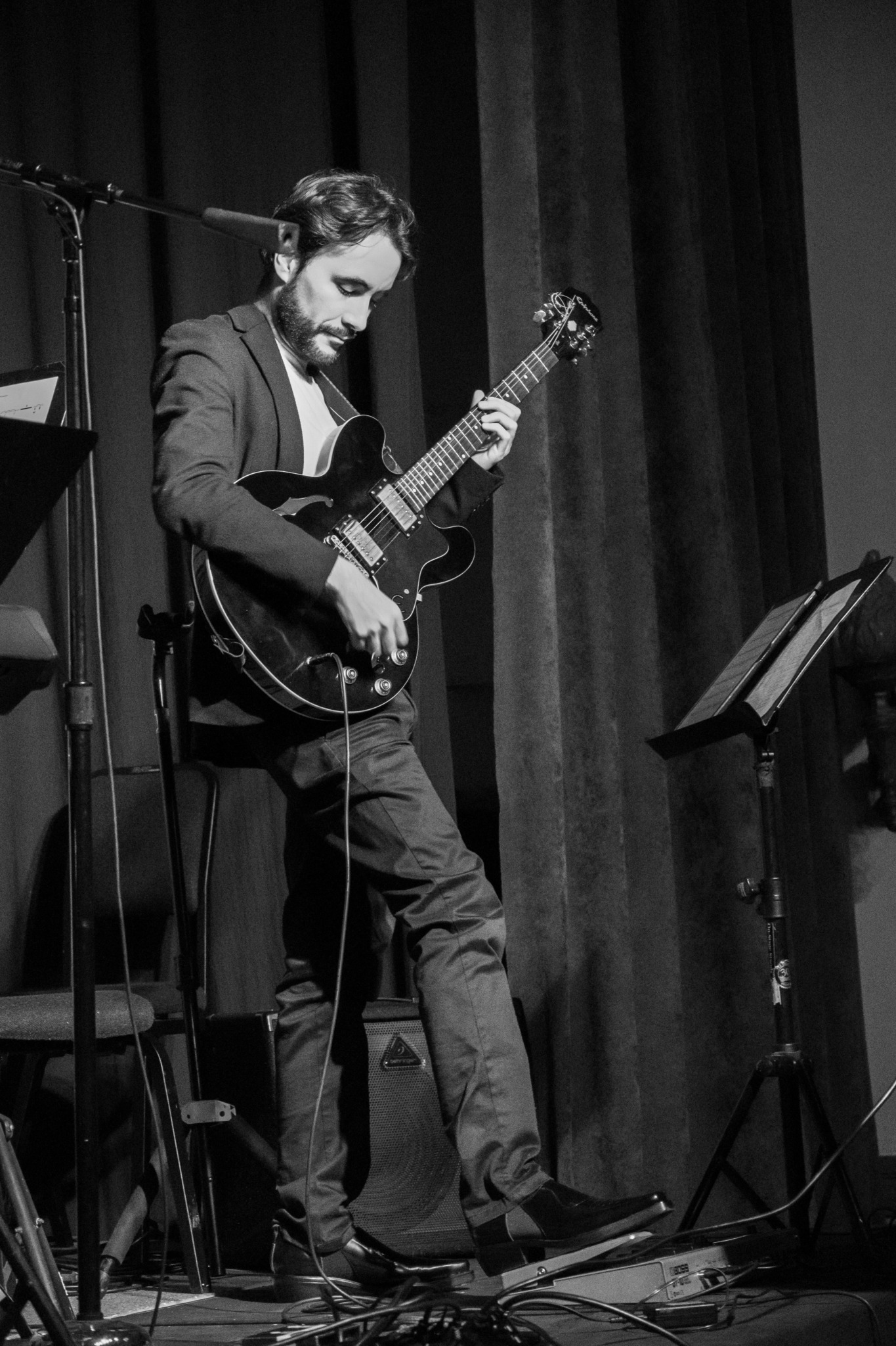 Man playing electric guitar on stage with music stand and curtains in background.
