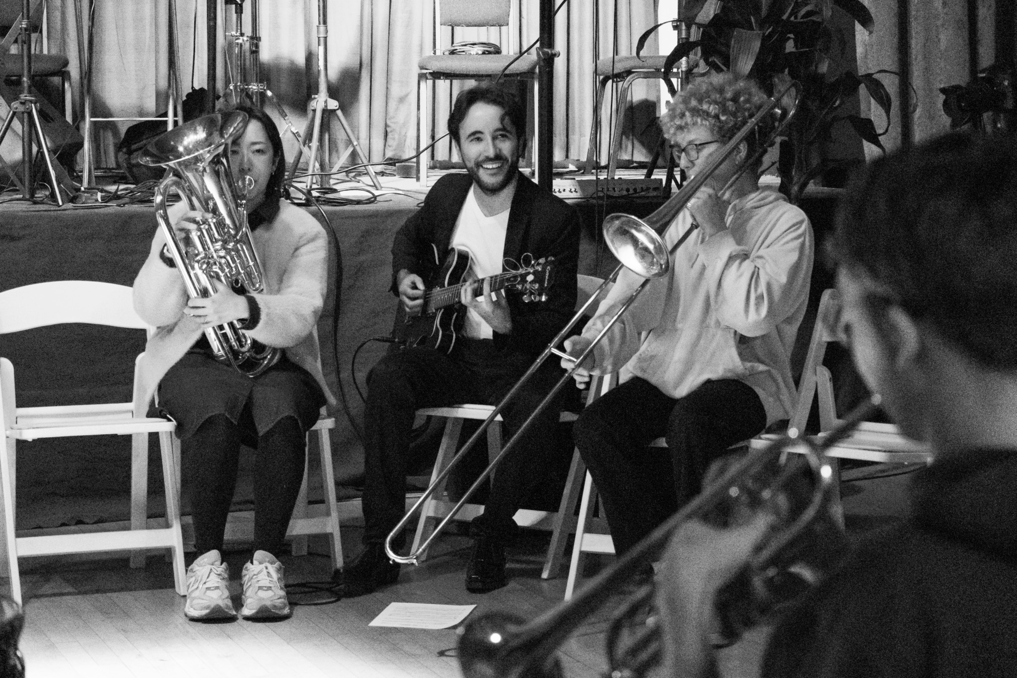 Three musicians seated with instruments, one person playing a trombone, in a rehearsal space with music stands and chairs.