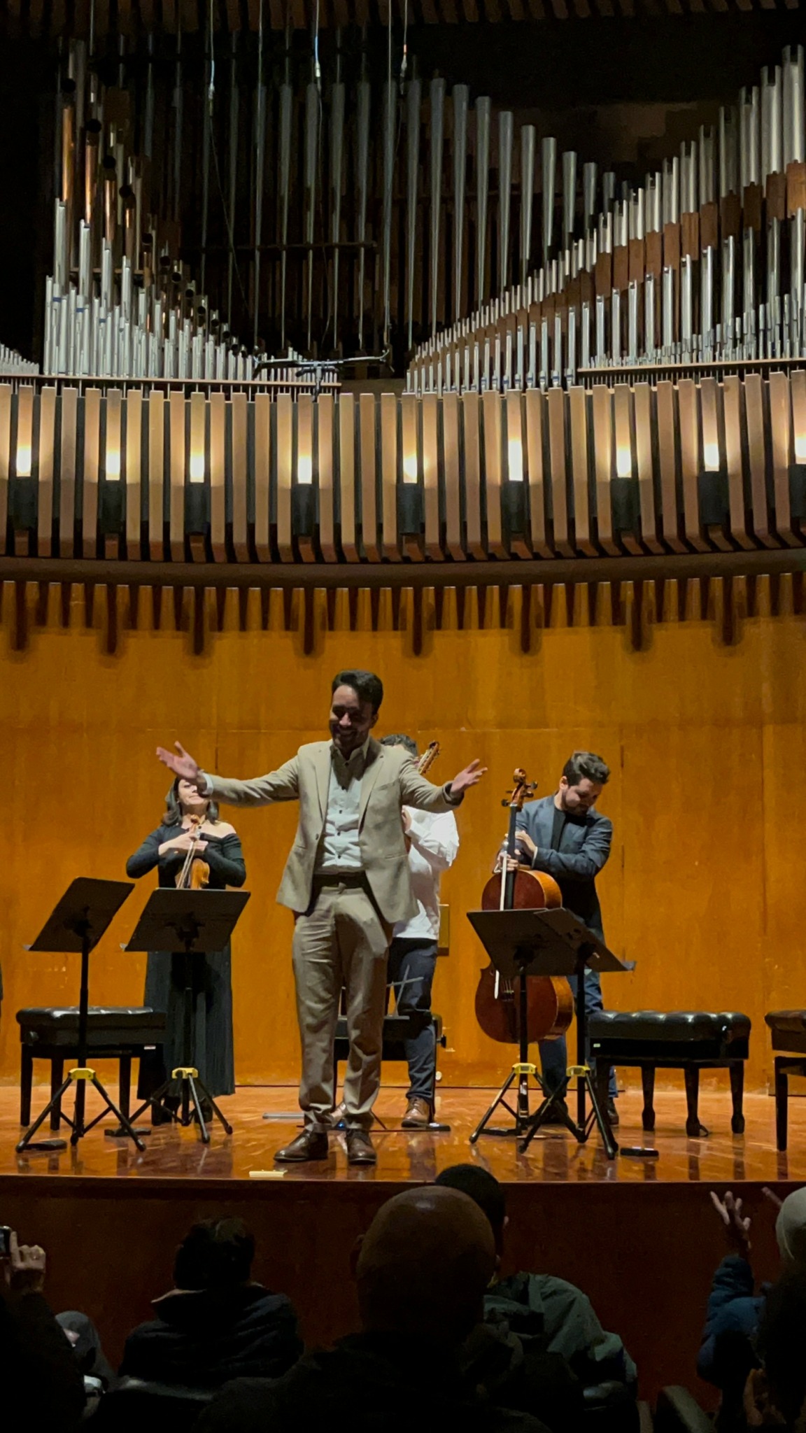 Three musicians on stage with instruments, one person standing and two seated, in a concert hall with pipes above.