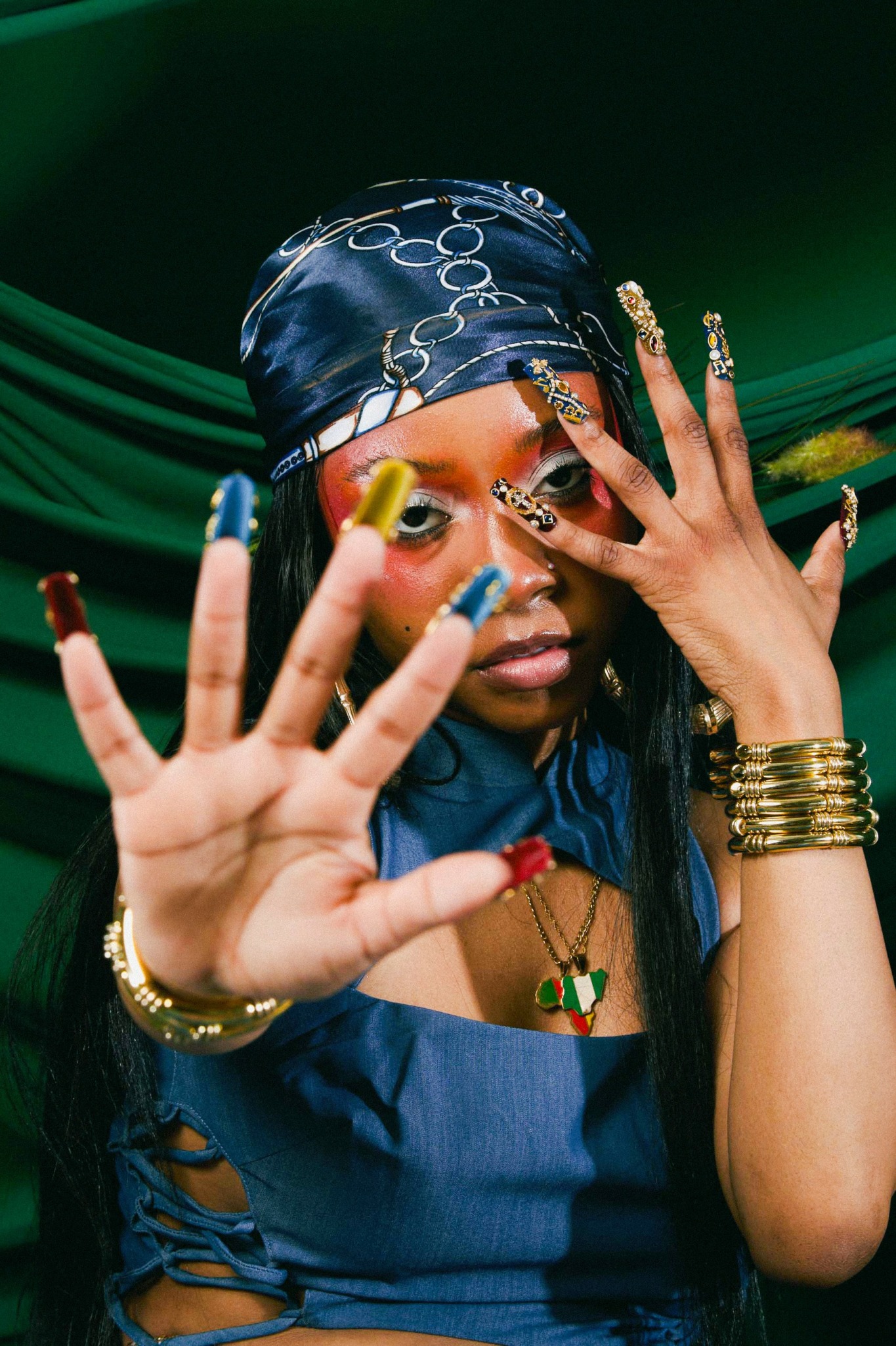 Person with colorful nails, jewelry, and a headscarf, holding hands up with fingers spread, against a green background.