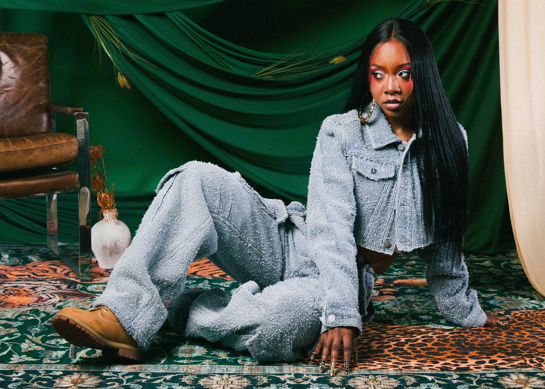 Woman with long black hair in gray outfit sitting on patterned carpet, green drapes in background.