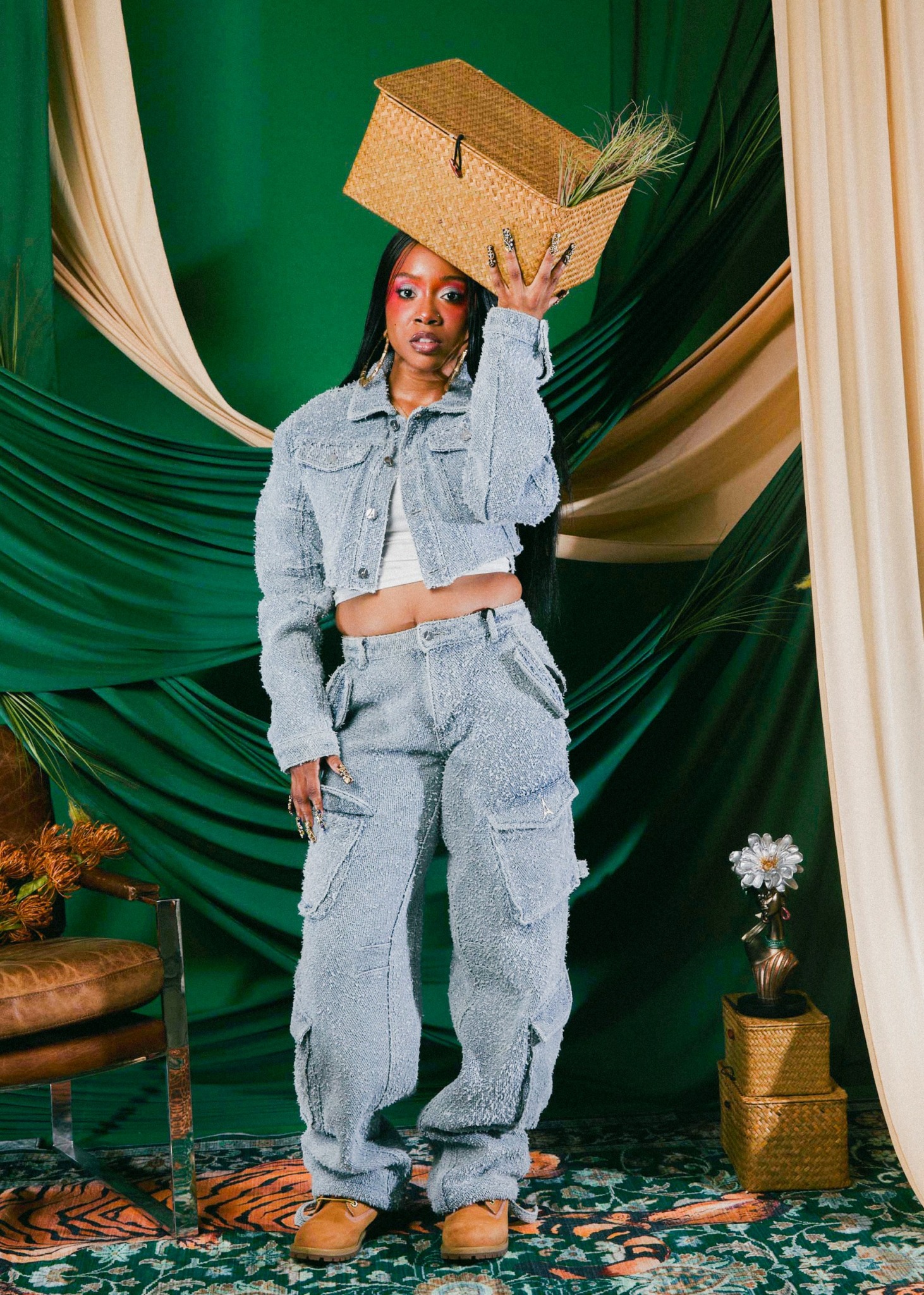 Woman in denim outfit balancing a woven basket on her head, standing in front of green and cream drapes.