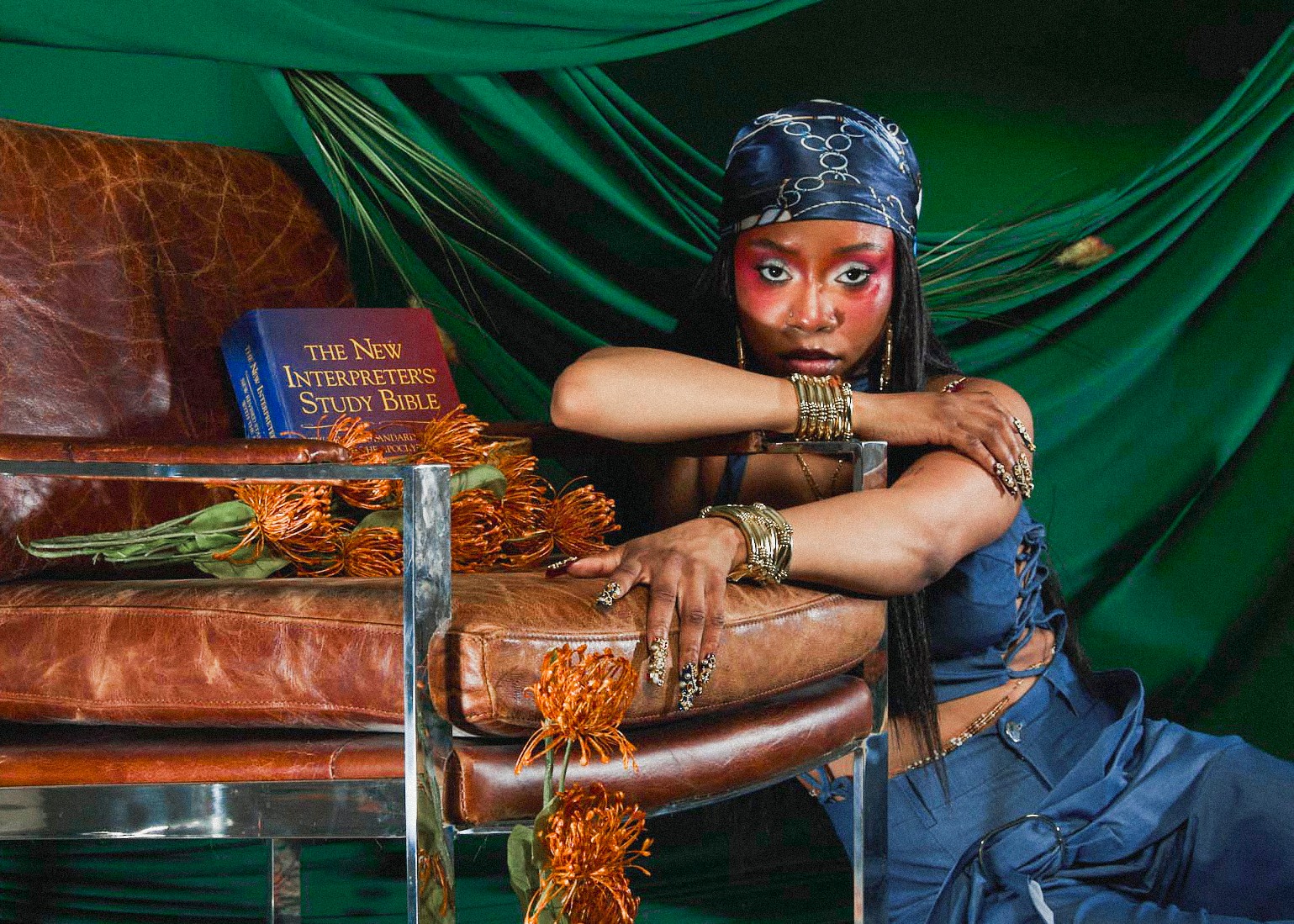 Woman with headscarf and jewelry leans on a table with a book and orange decorations, green drapes in background.