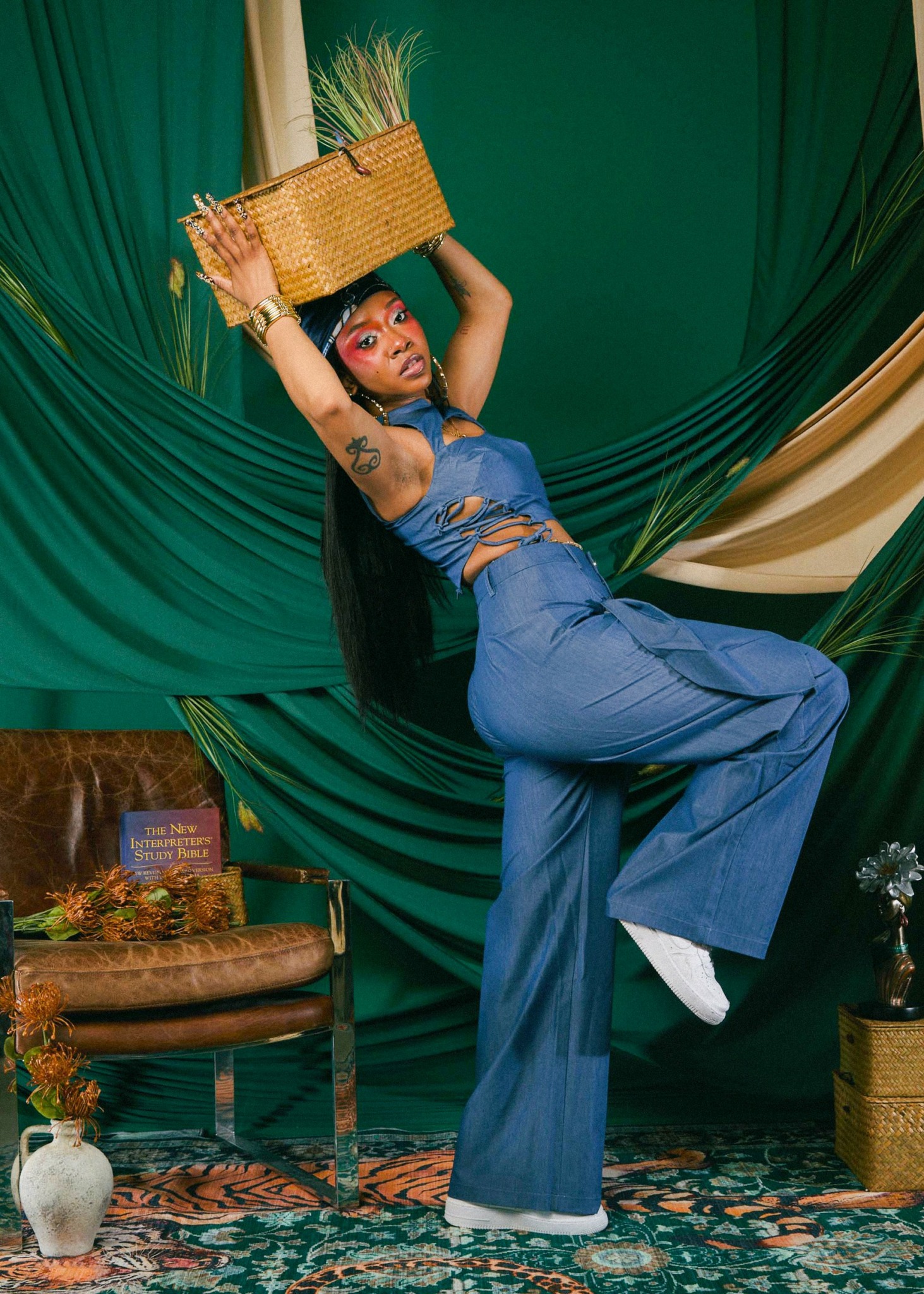 Person with a basket on their head, standing on one leg, in front of green and beige curtains.