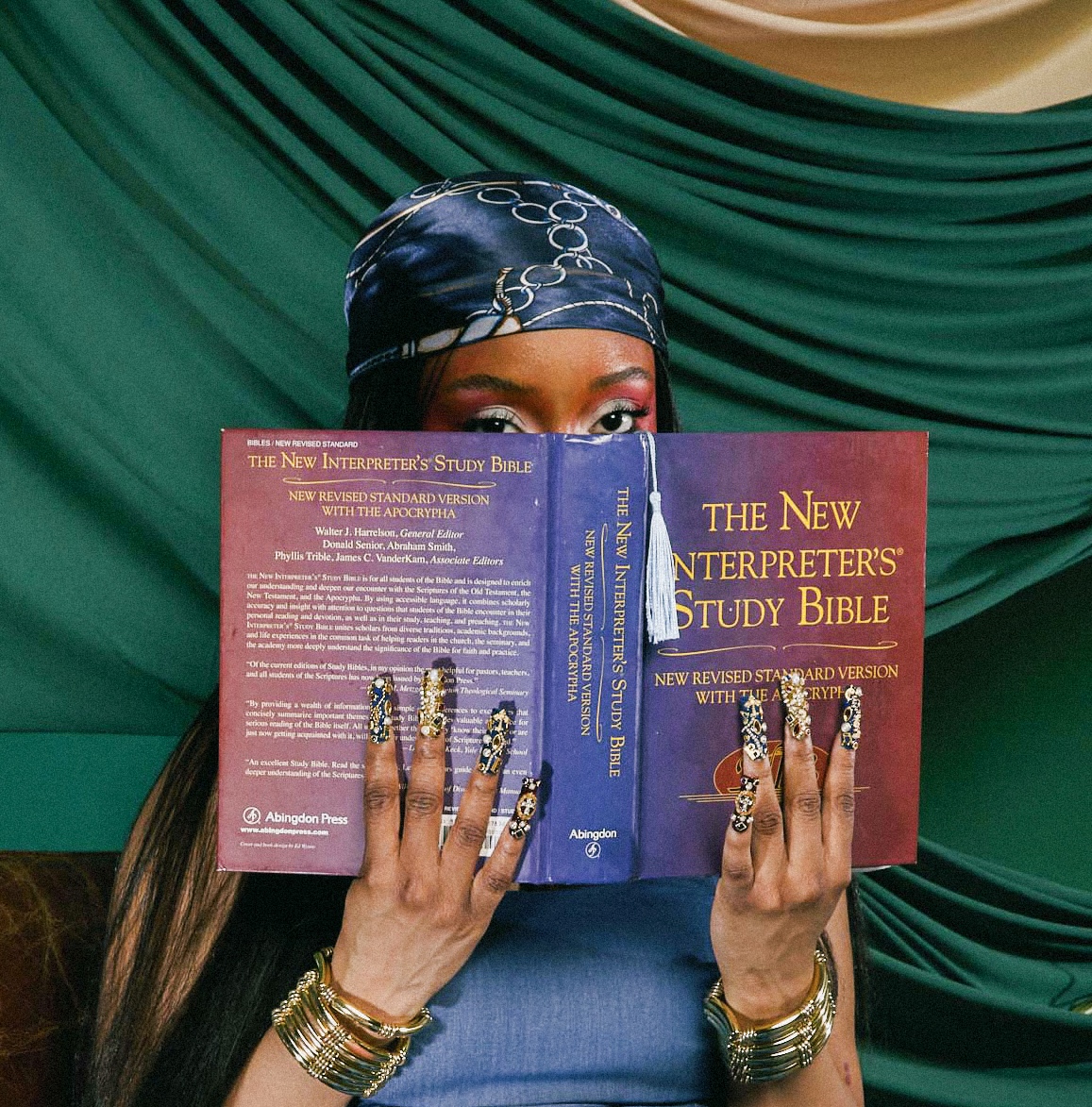 Person with headscarf reading a Bible titled 'The New Interpreter's Study Bible' with a green draped background.
