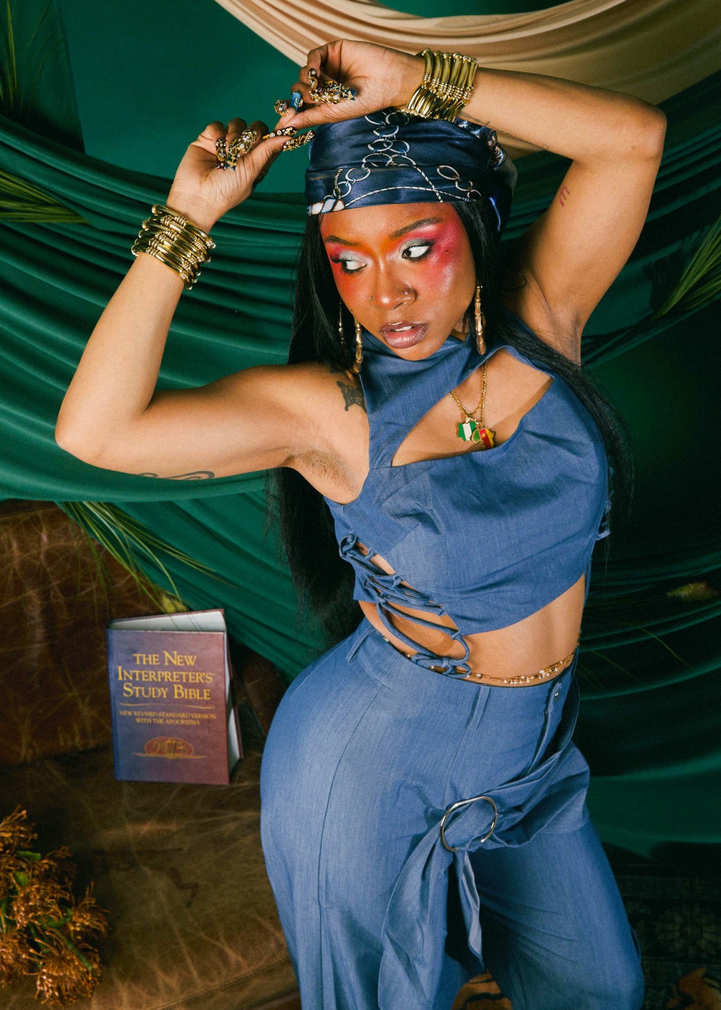 Woman with long dark hair, wearing a blue bandana, sleeveless top, and jewelry, poses with arms raised.