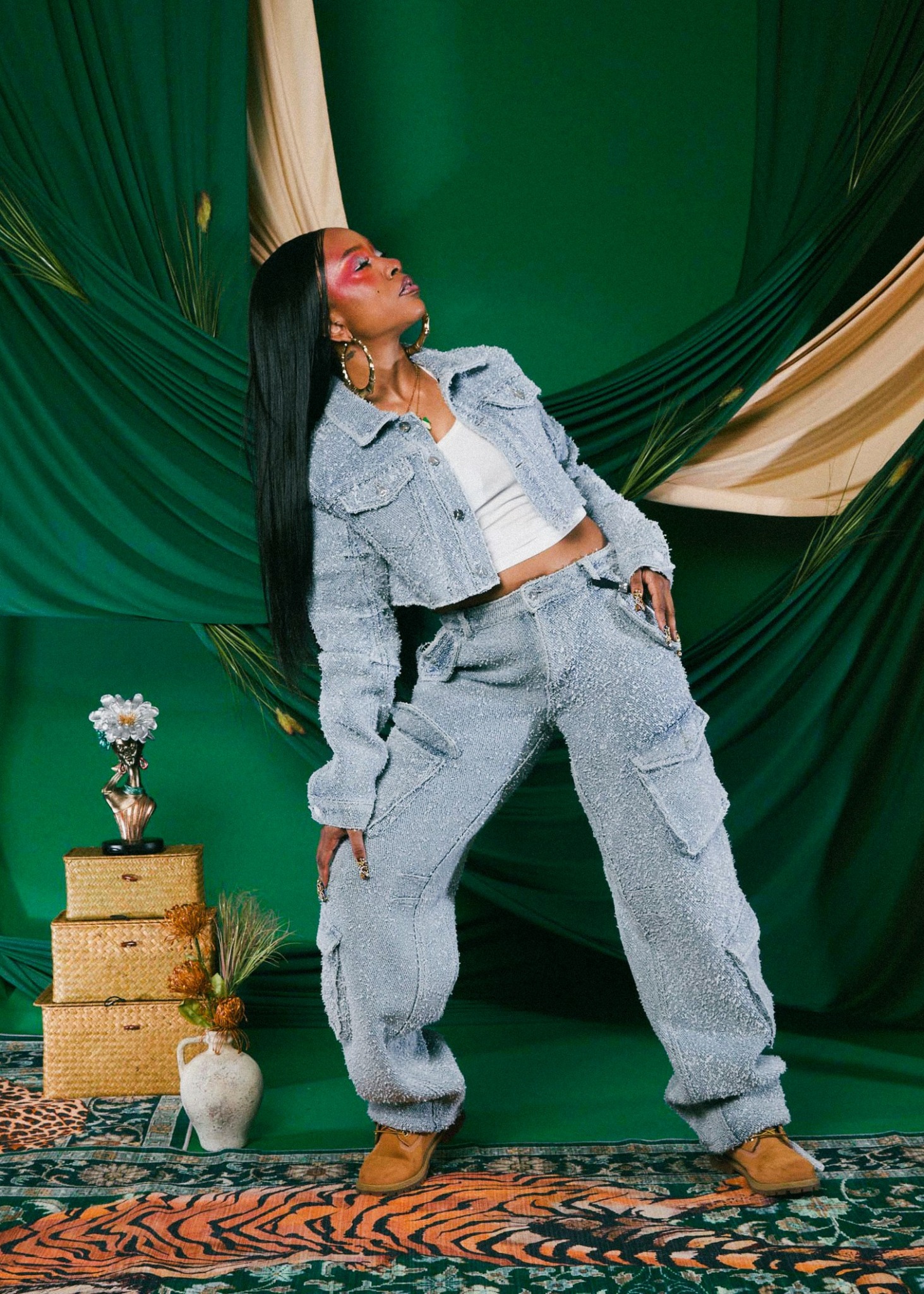 Woman with long dark hair in a gray outfit poses in front of green and beige drapes, with a small table and flowers nearby.