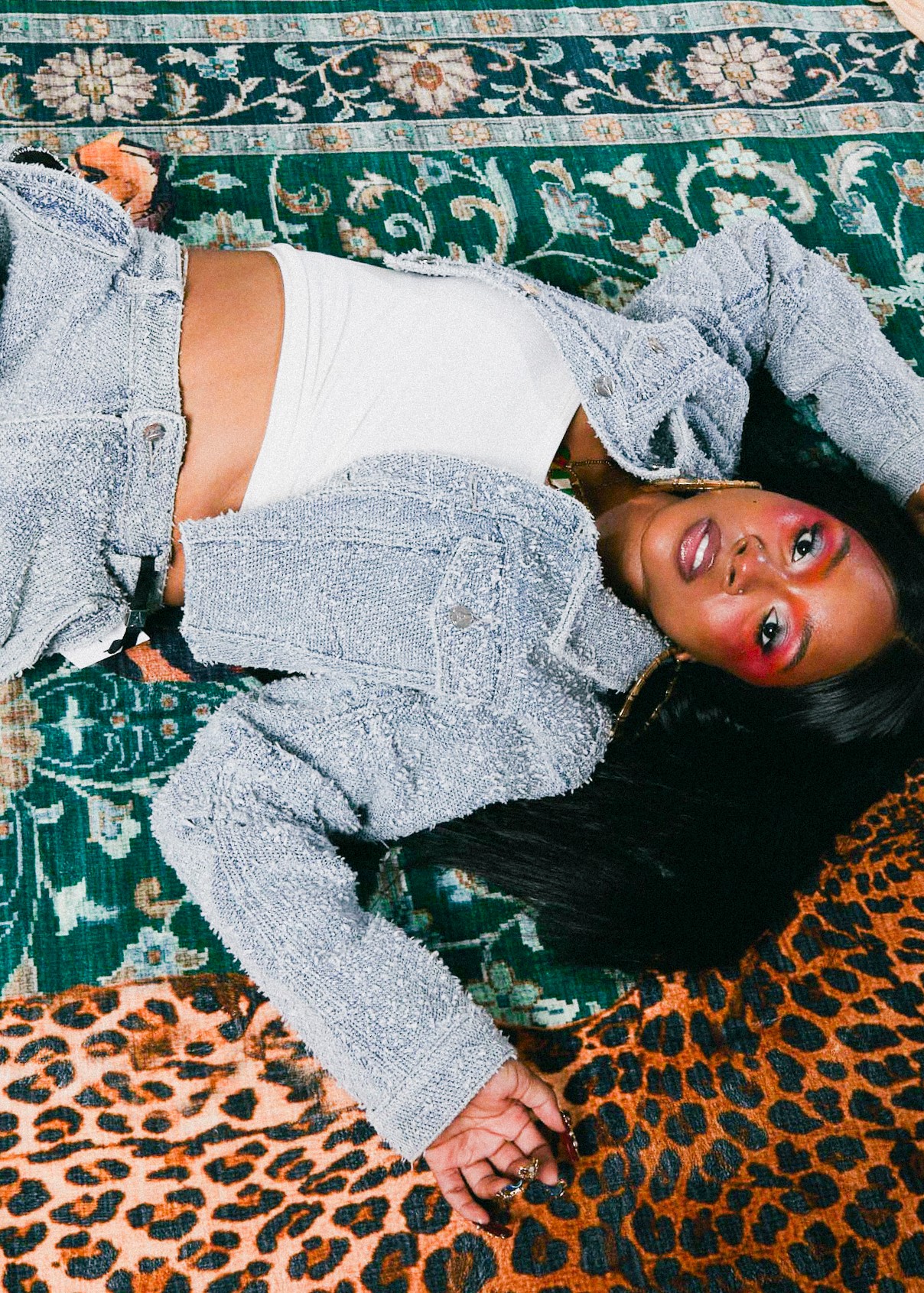 Woman lying on patterned carpet, wearing a gray jacket and white top, with long dark hair and makeup.