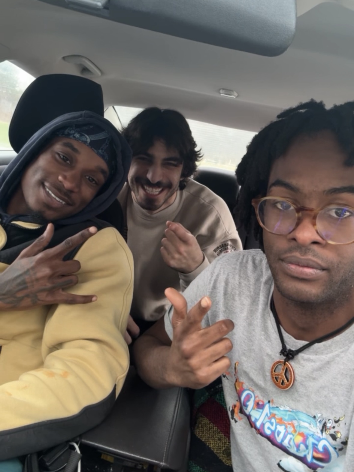 Three people sitting in a car, smiling and making hand gestures, with a light-colored interior and headrests visible.