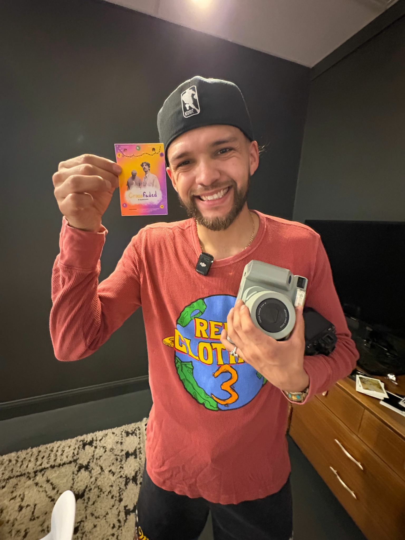 Person smiling, holding a colorful card in one hand and a camera in the other, wearing a red shirt and black cap.