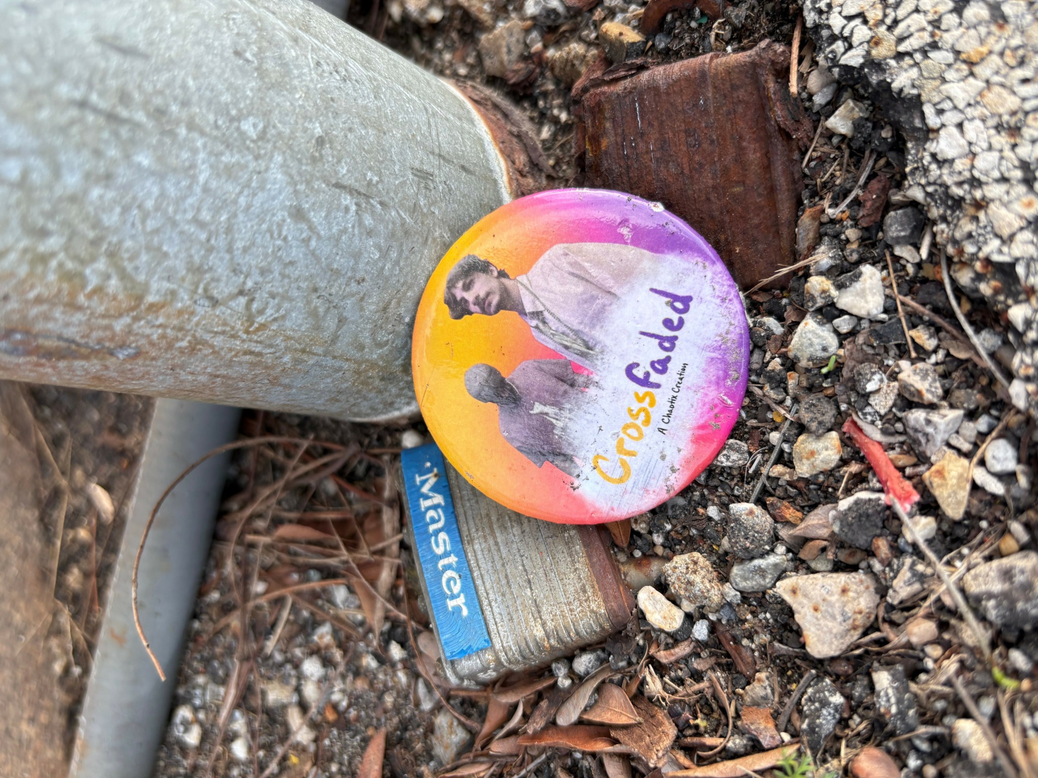 Colorful button with a person and the words 'Cross Faded' on the ground among dirt and small rocks.
