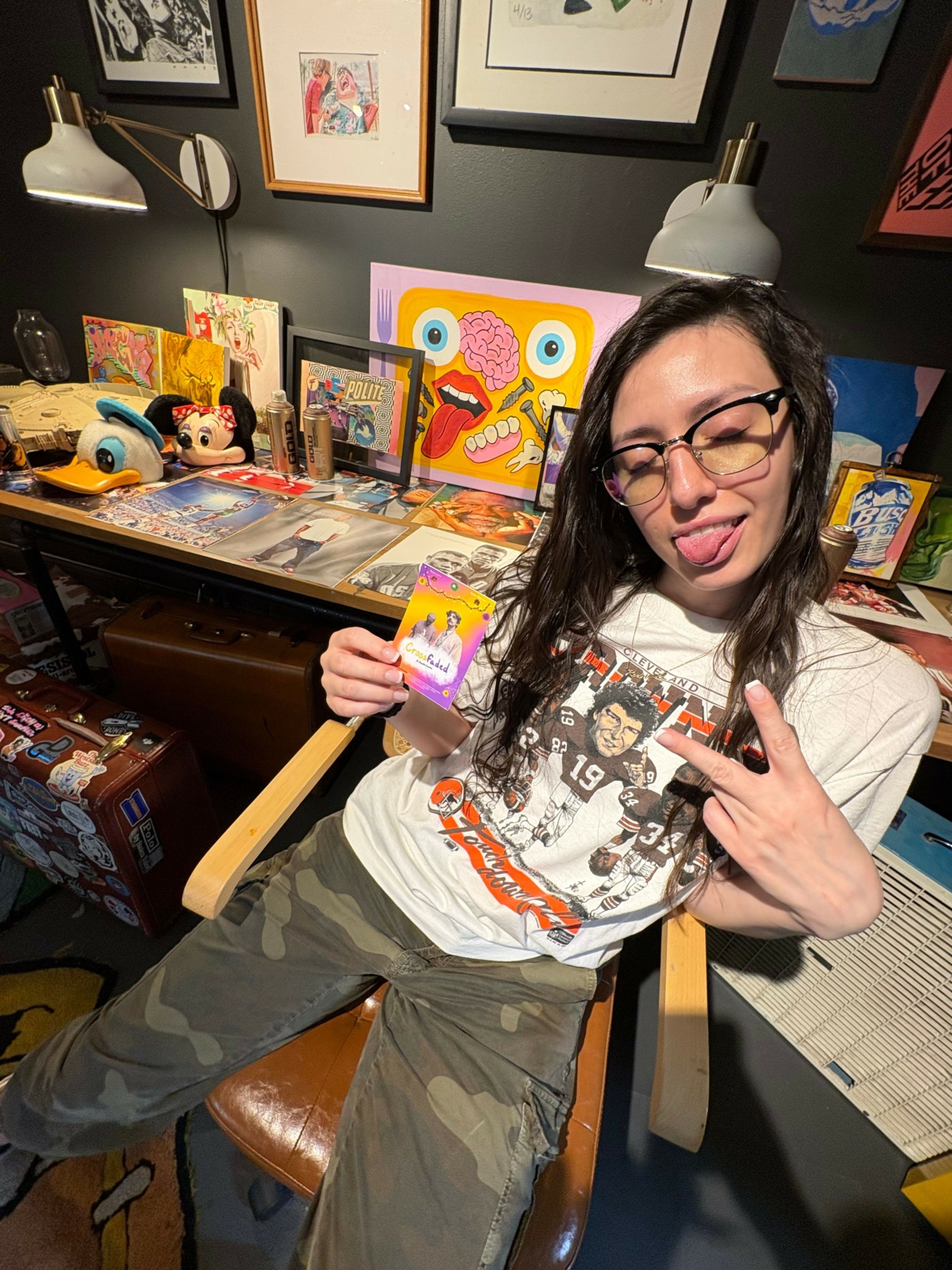 Person with glasses making a peace sign, sitting in a room with colorful artwork and collectibles on shelves.