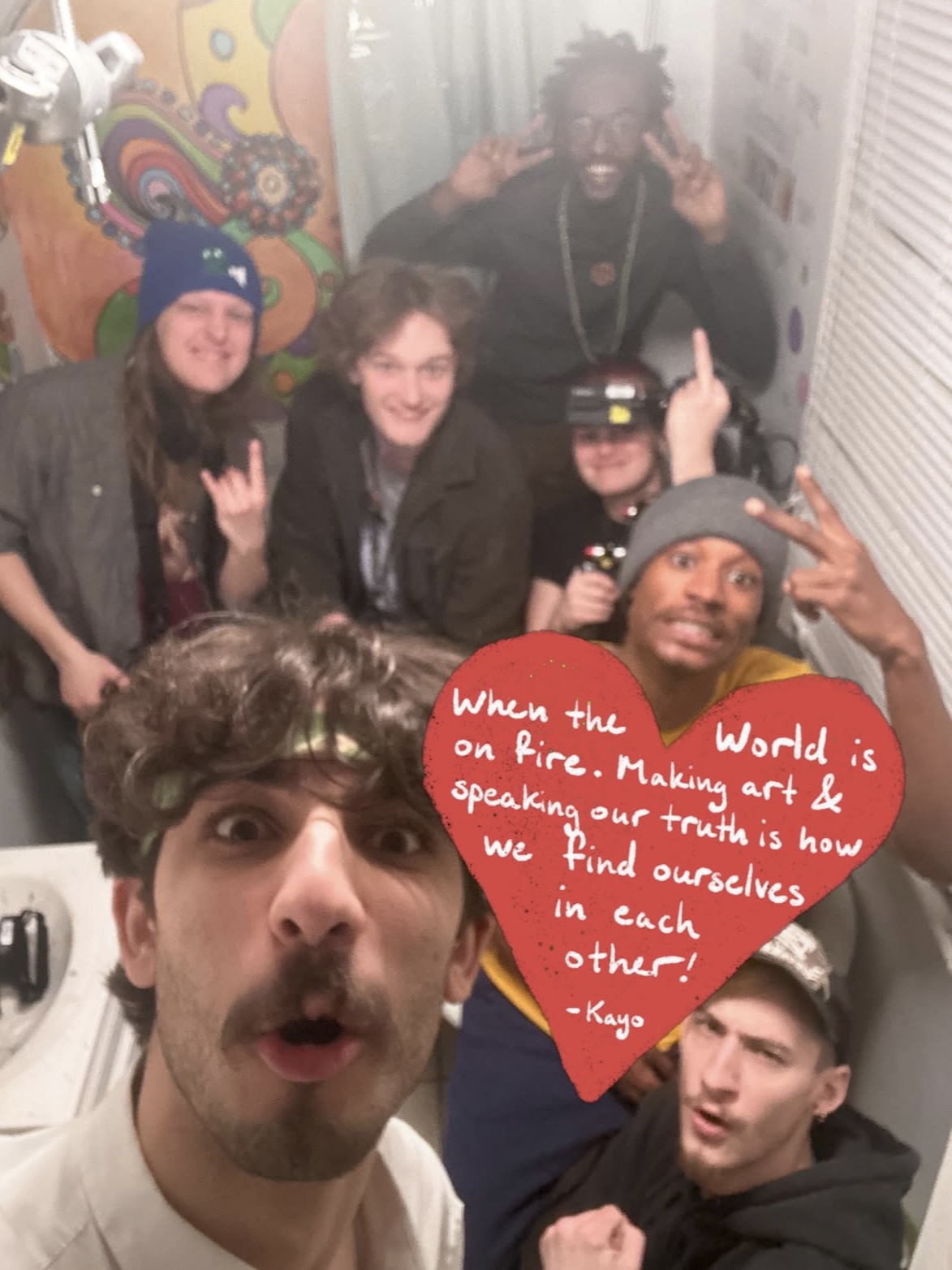 Group of six people indoors, some making hand gestures, one holding a red heart-shaped sign with a quote, smiling and posing.