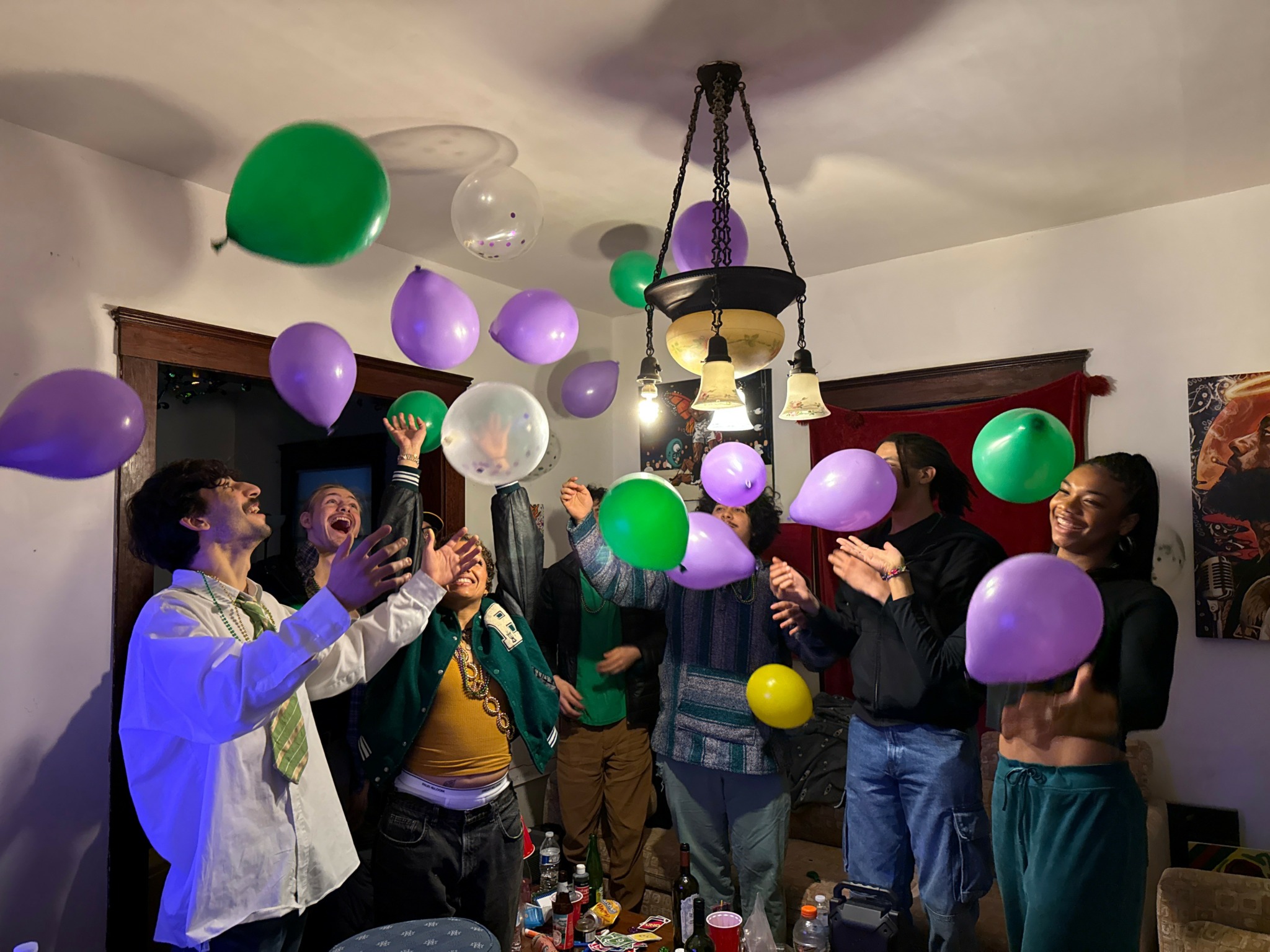 Group of people celebrating indoors with colorful balloons, some reaching for balloons, in a room with a chandelier and artwork.