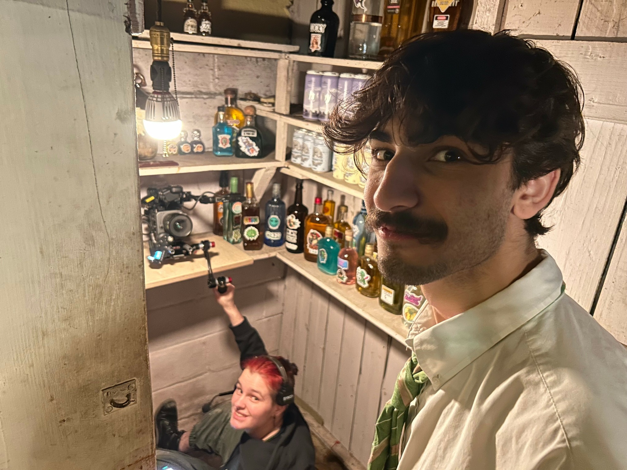 Man with dark curly hair and mustache in foreground, smiling woman with red hair in background, shelves with bottles behind them.