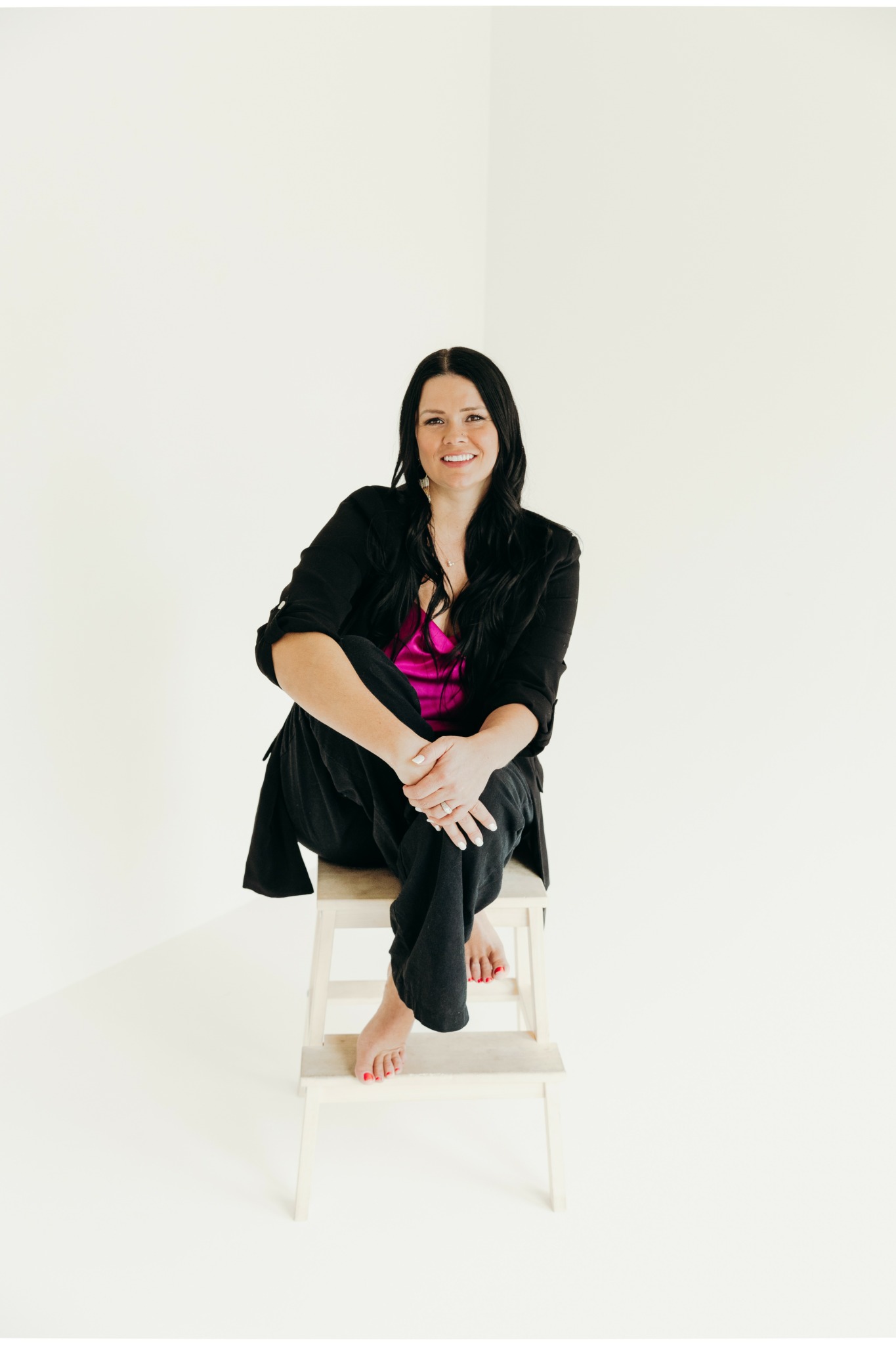 Woman sitting on a stool with crossed legs, smiling, wearing a black blazer and pink top, in a white room.