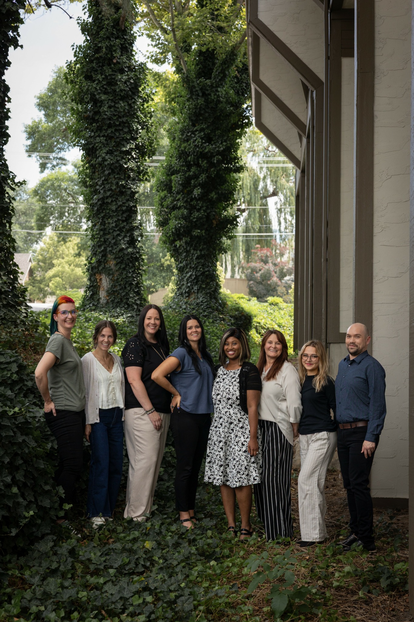 Group of nine people standing outdoors in front of trees and a building, smiling at the camera.