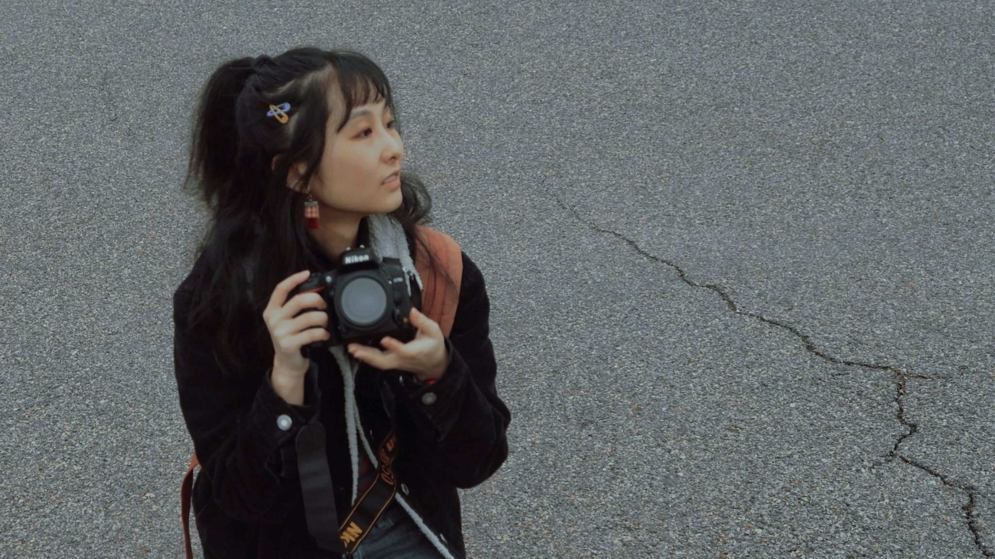 Young woman holding a camera, standing on cracked asphalt ground, looking to the side.