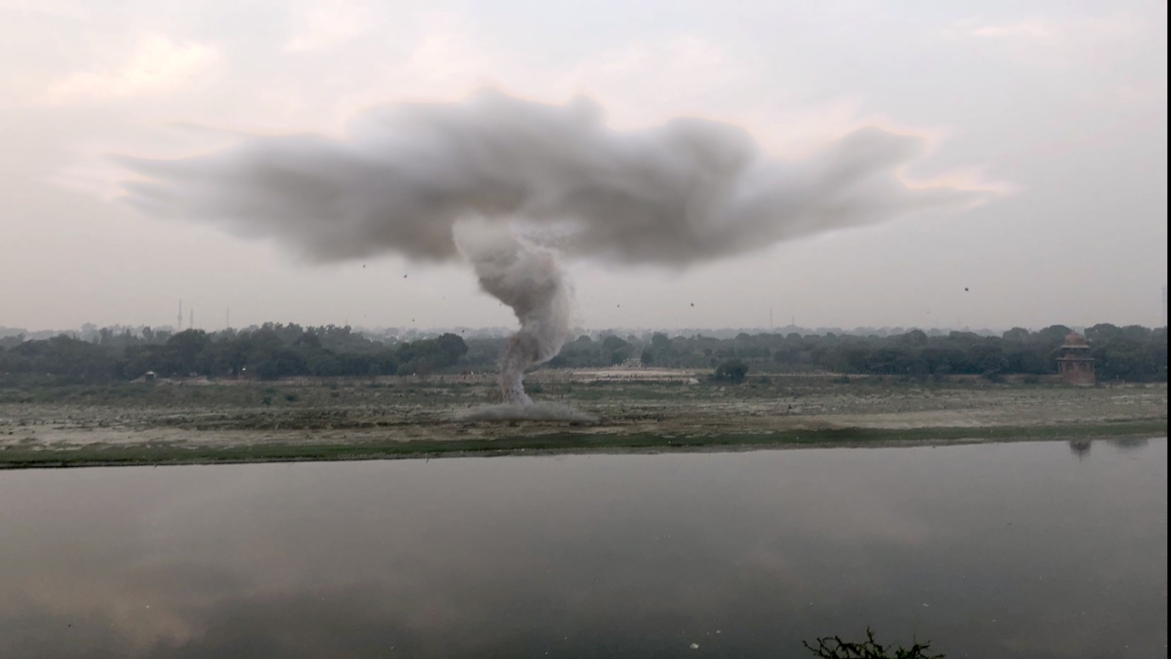 Smoke rising from a fire near a distant treeline, with a body of water in the foreground.