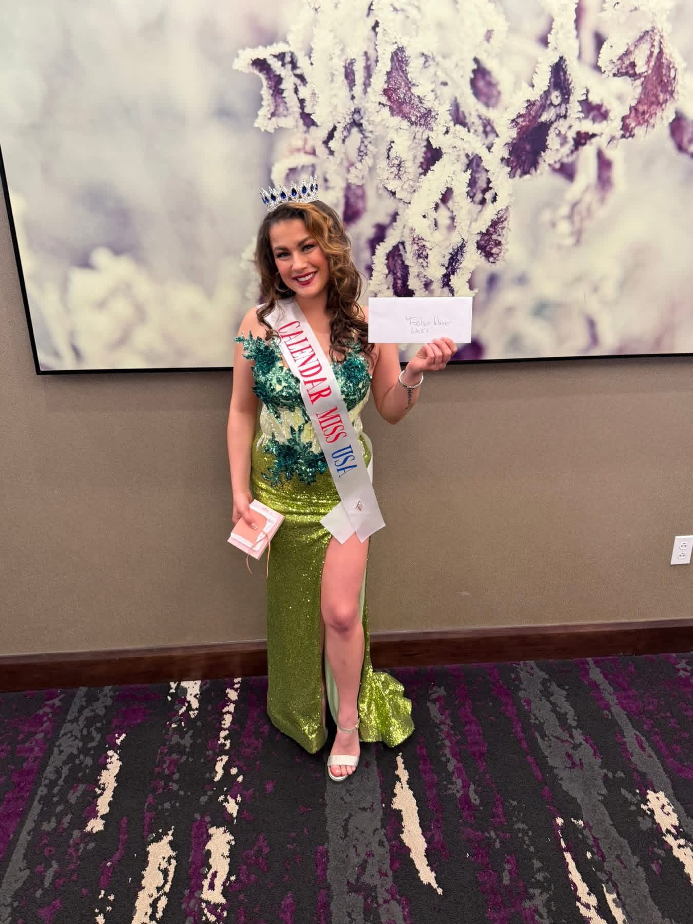 Young woman in a green dress with a sash, holding a certificate, standing in front of abstract art.