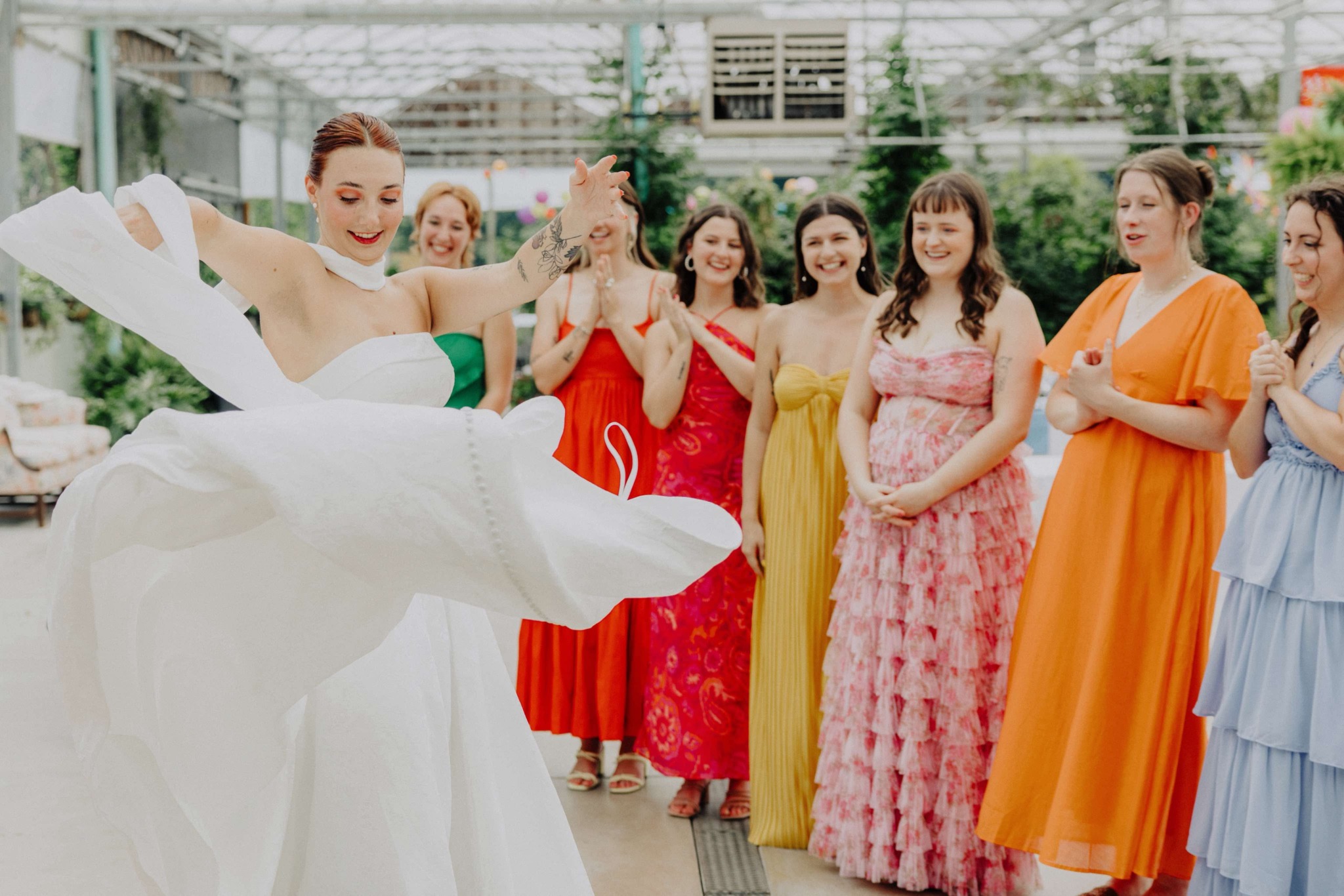 A bride in a white wedding dress twirls with a group of women in colorful dresses inside a greenhouse.