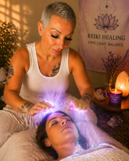 Woman with short gray hair performs Reiki healing on a woman lying down with closed eyes in a dimly lit room.