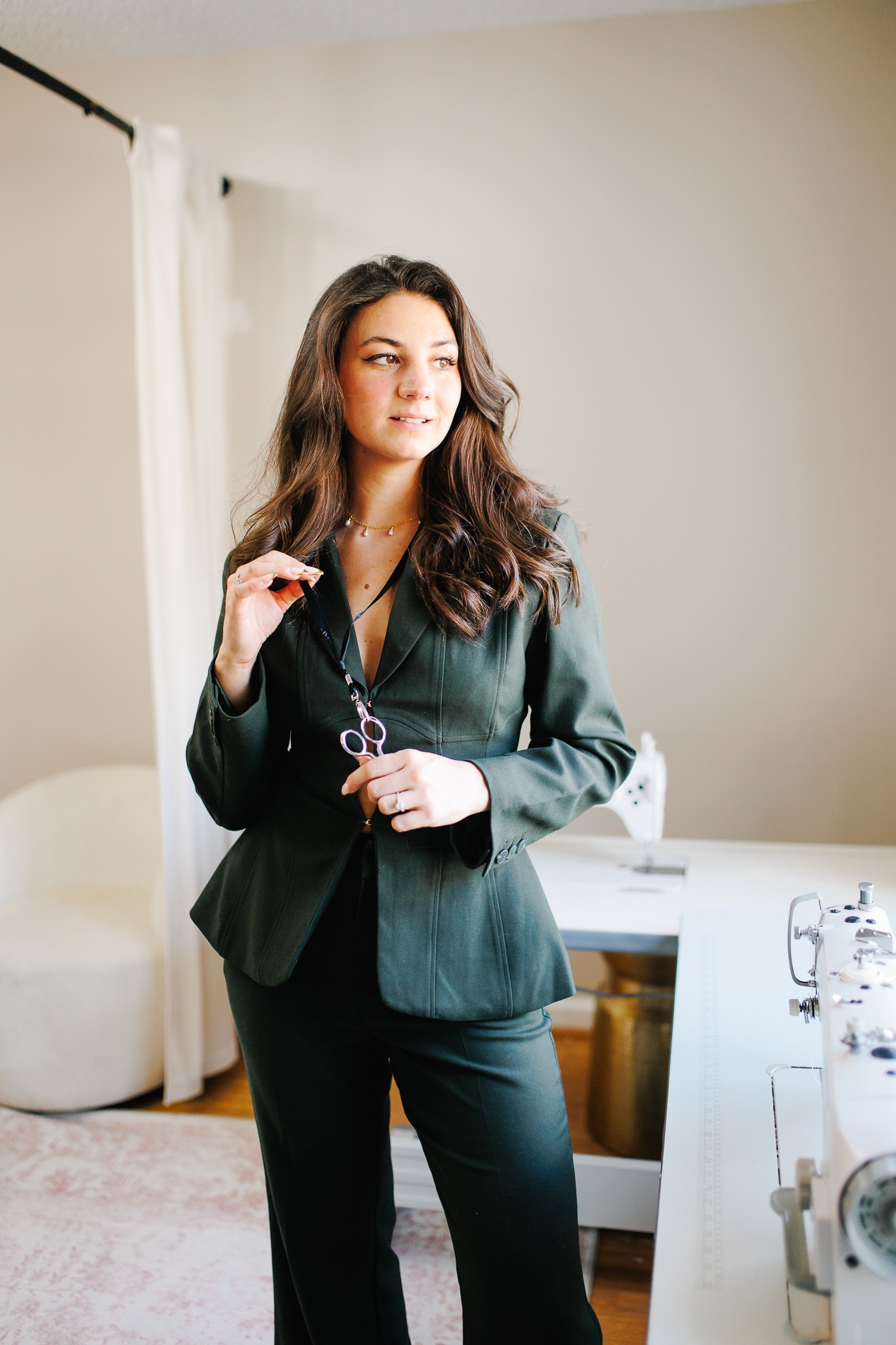 Woman with wavy hair in a dark blazer holding glasses, standing in a room with white walls and furniture.