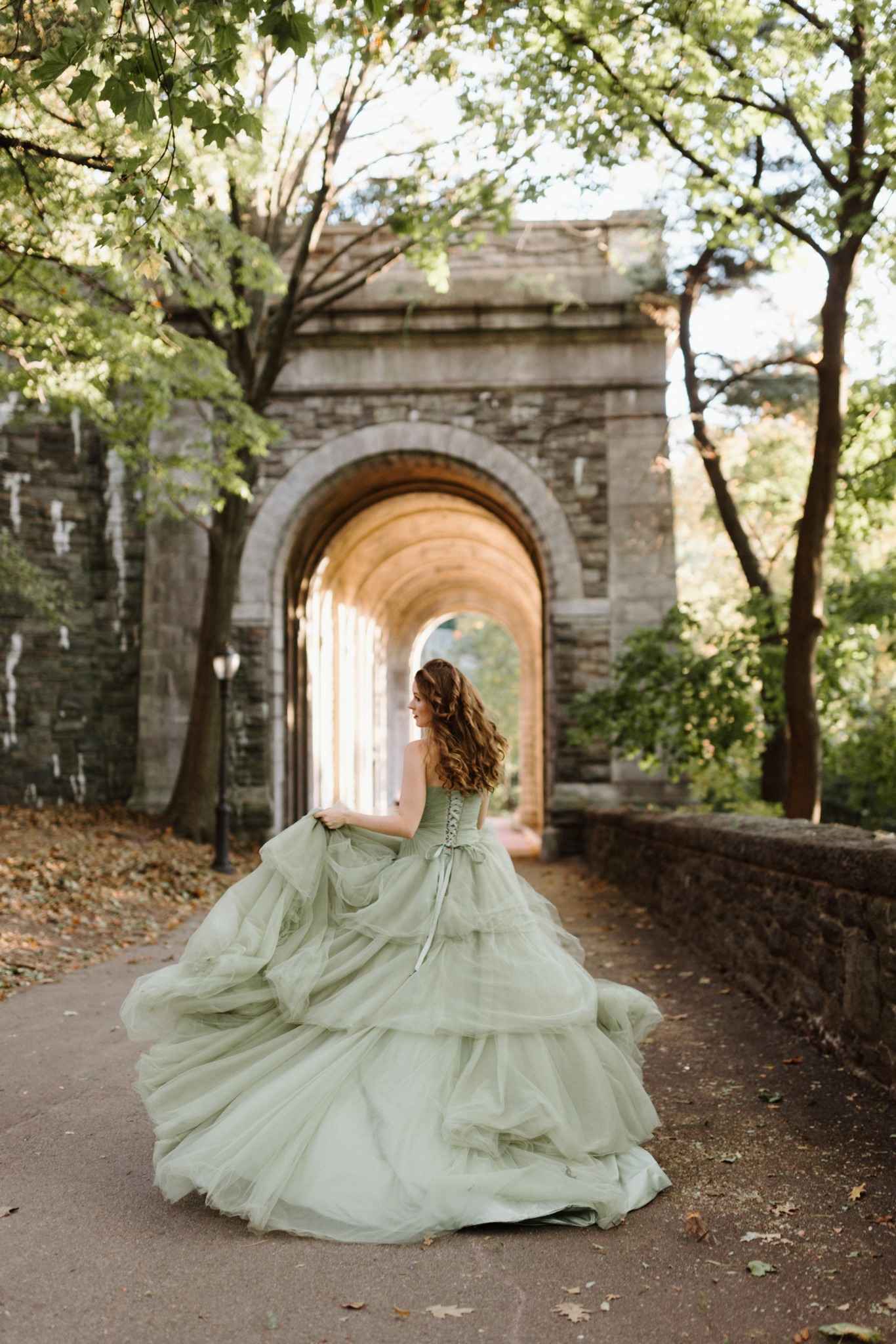 Woman in a flowing light green dress sitting on a path near an arched stone gateway surrounded by trees.