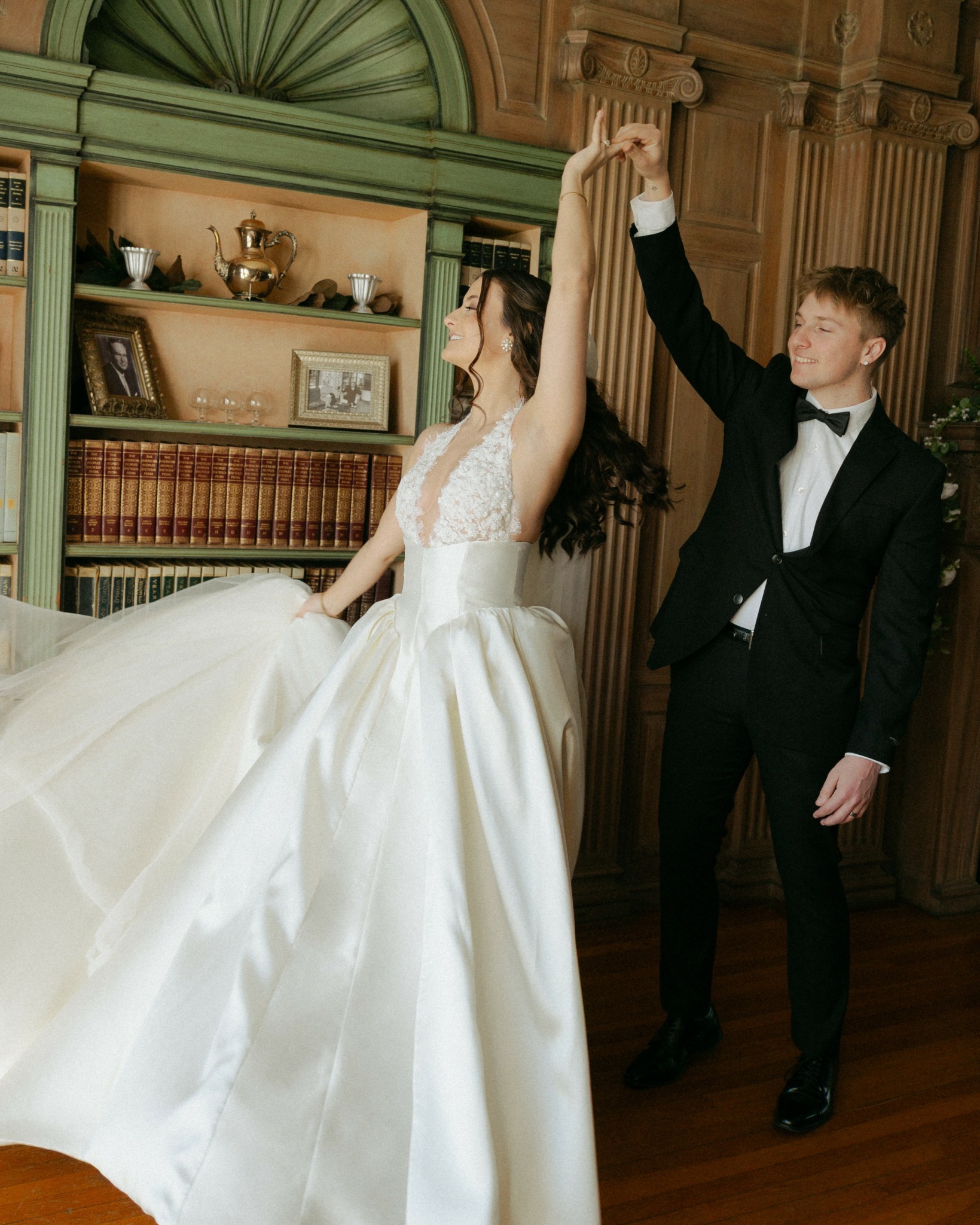 Bride and groom dancing indoors, holding hands, with wooden shelves and framed photos in background.