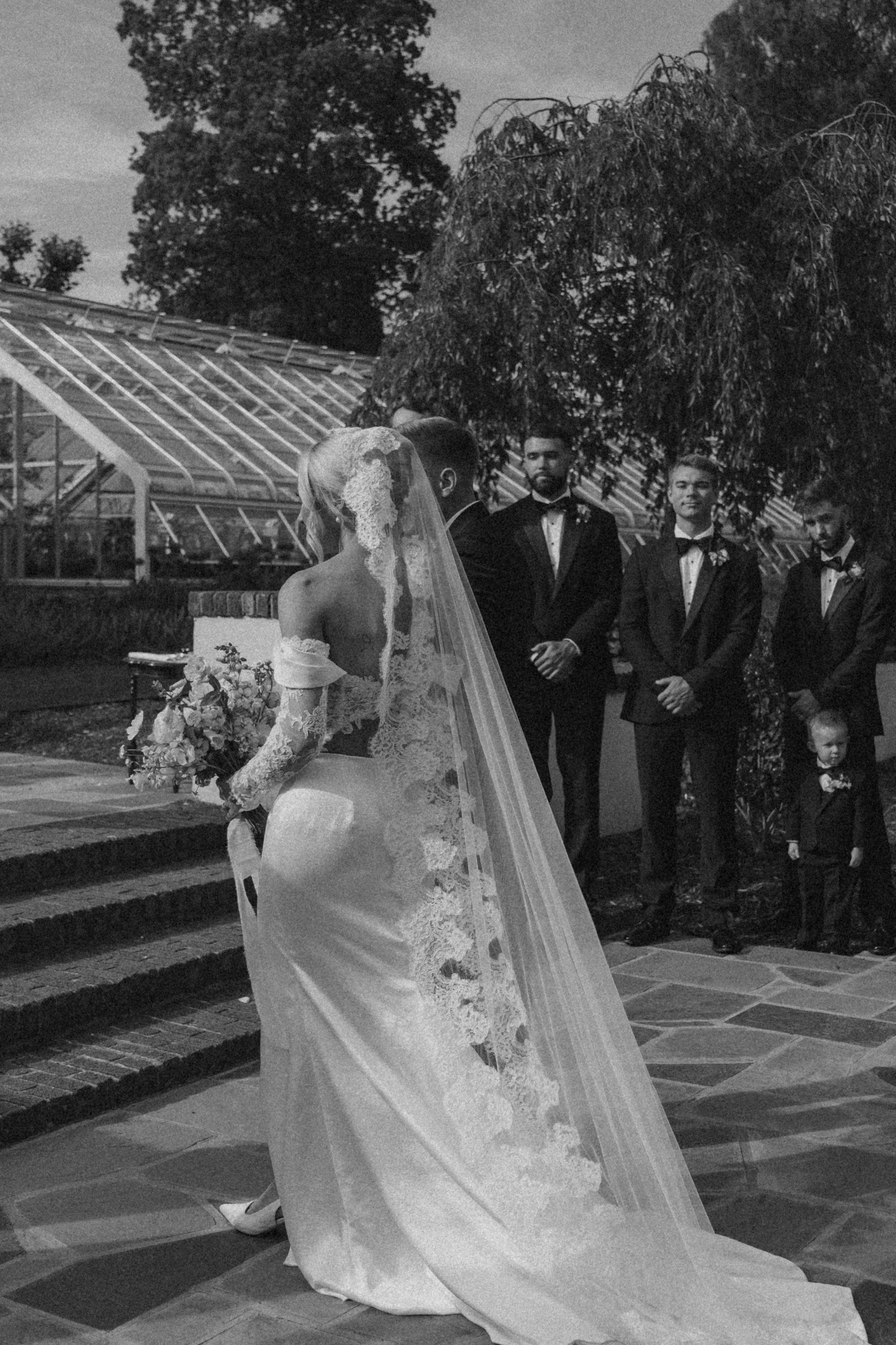 Bride in wedding dress and veil standing outdoors, with guests in formal attire nearby, trees and greenhouse in background.