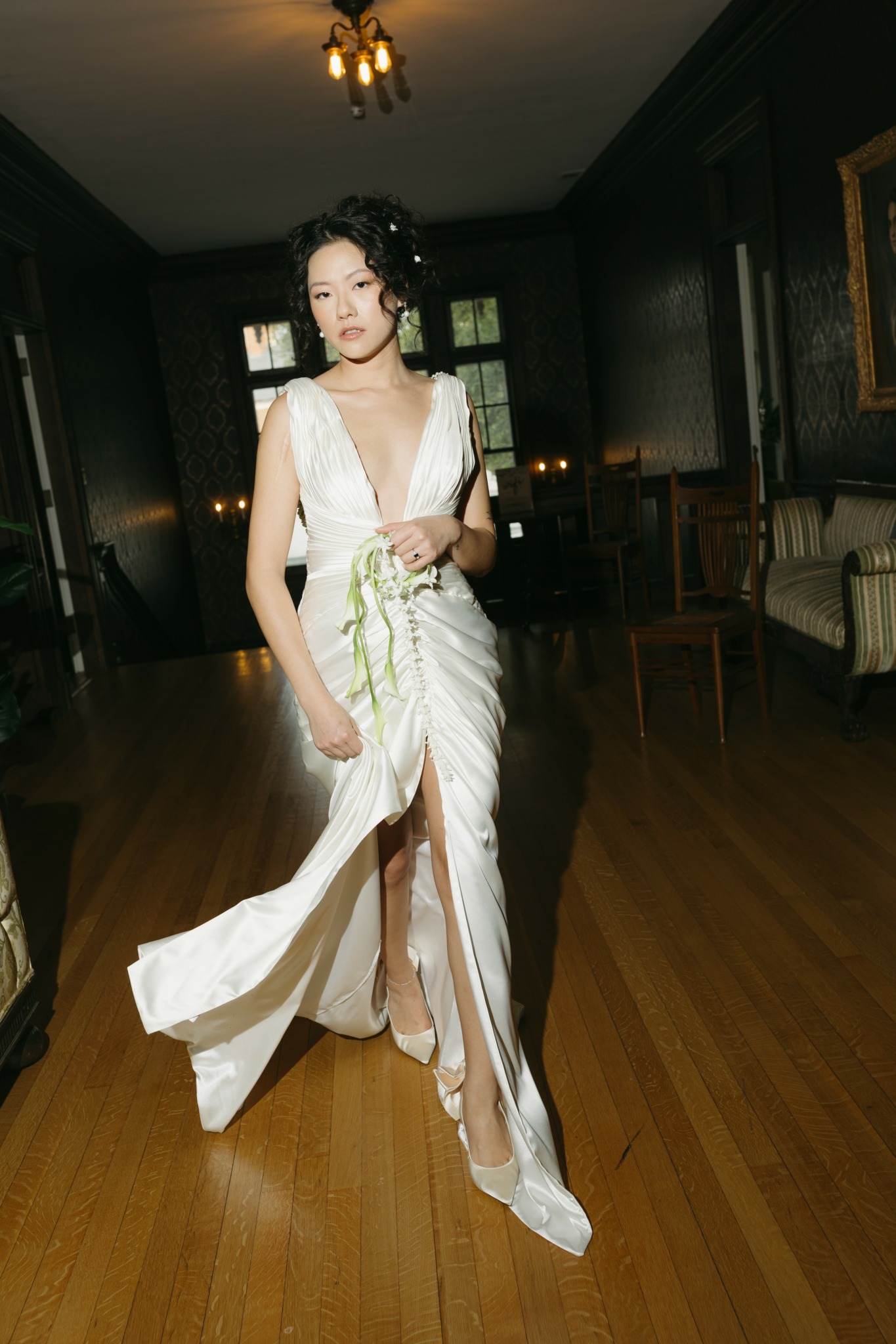 Young woman in a white dress with a slit, standing in a dimly lit room with wooden floors and dark walls.