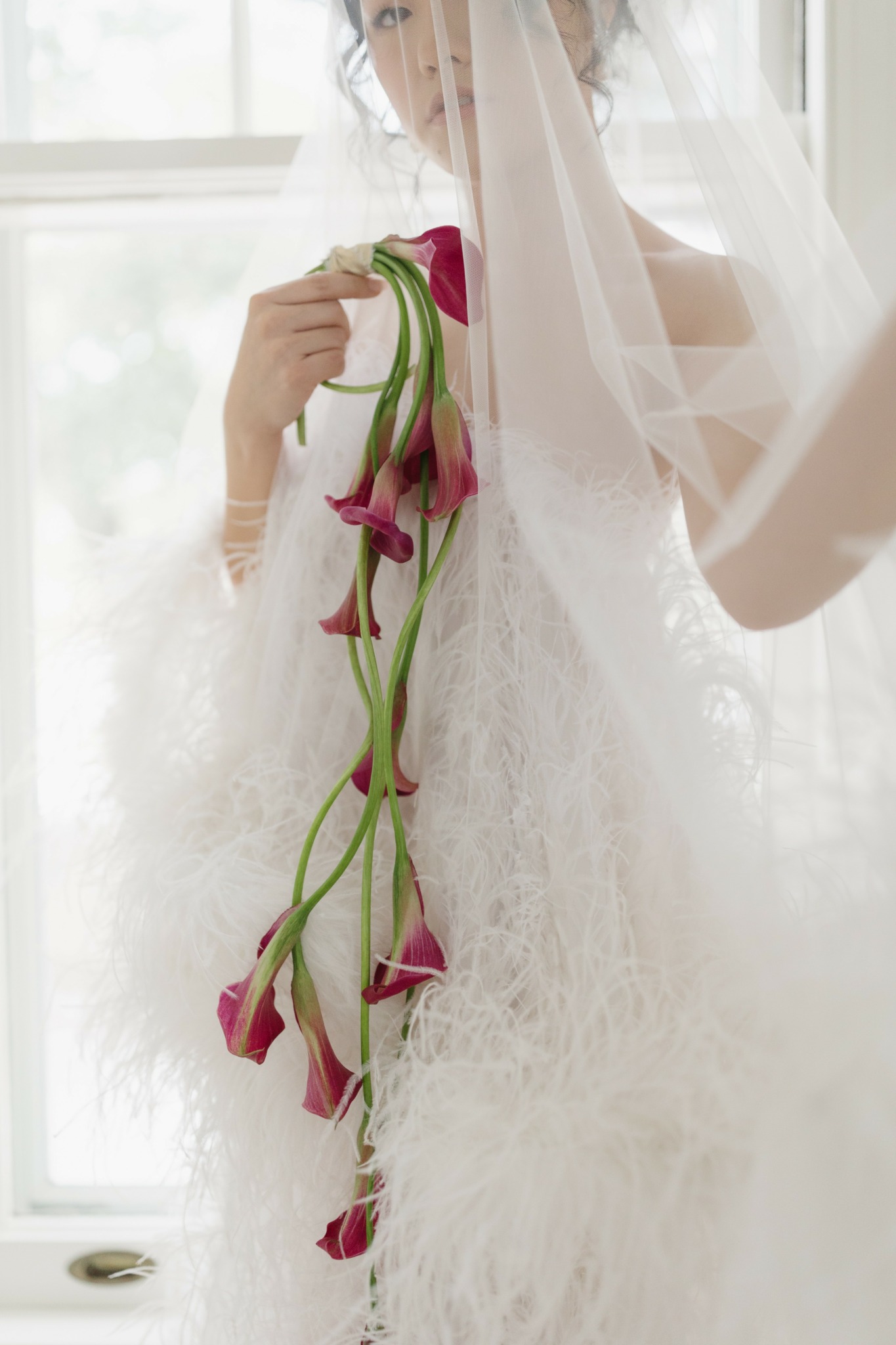 Person in wedding dress holding a bouquet of pink calla lilies behind a sheer curtain.