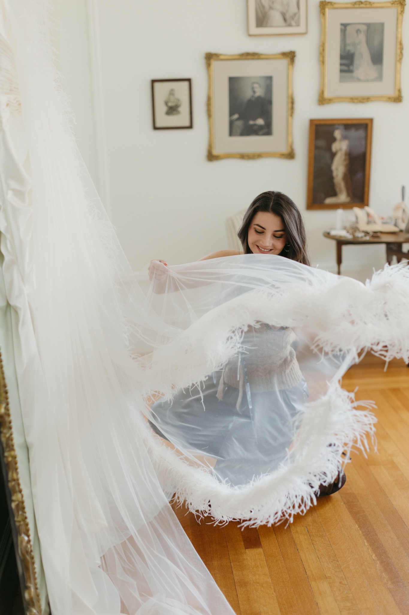 Young woman with dark hair holding a large white lace fabric in a room with framed pictures on the wall.