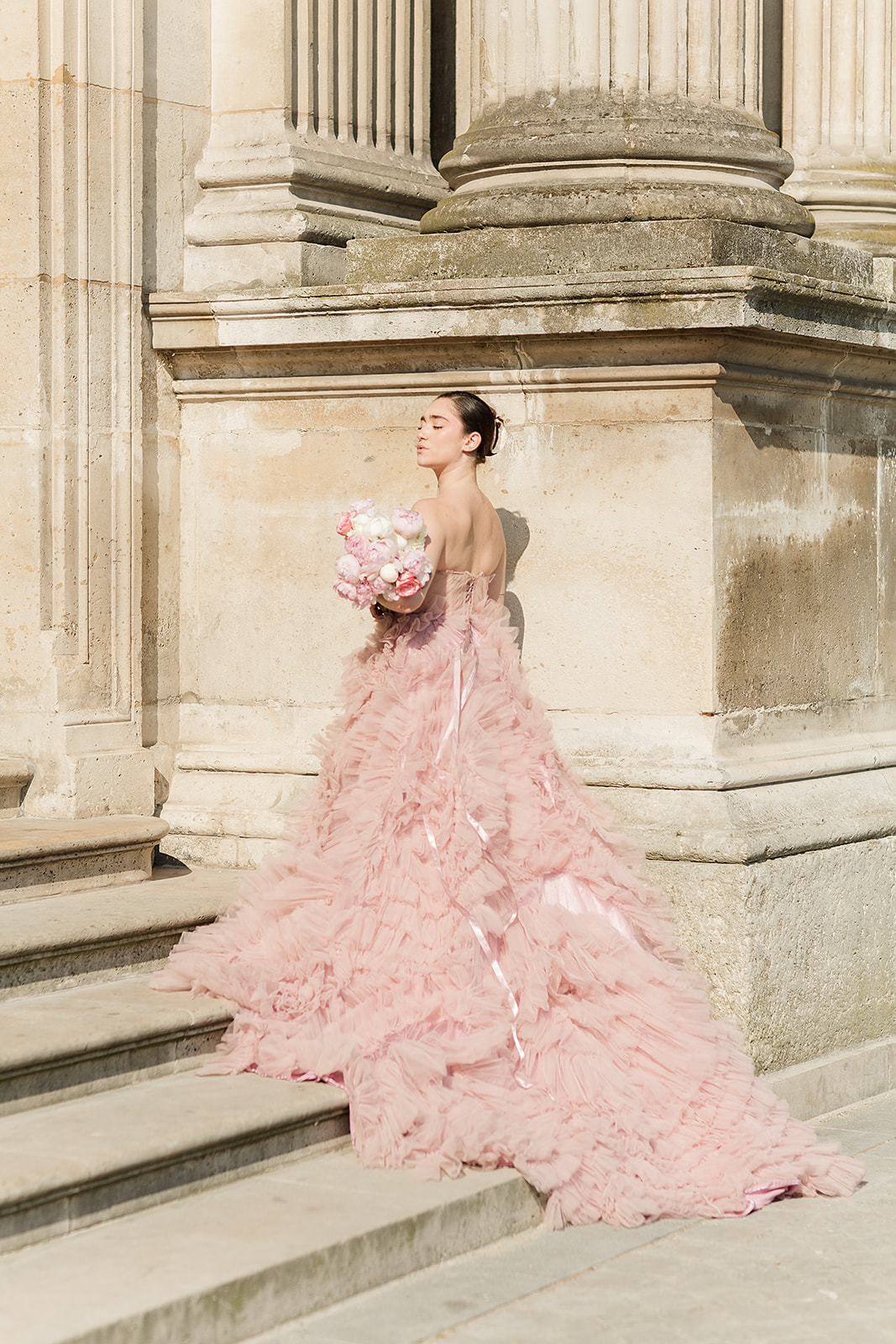 Woman in pink gown holding bouquet standing on stone steps outside a classical building with large columns.