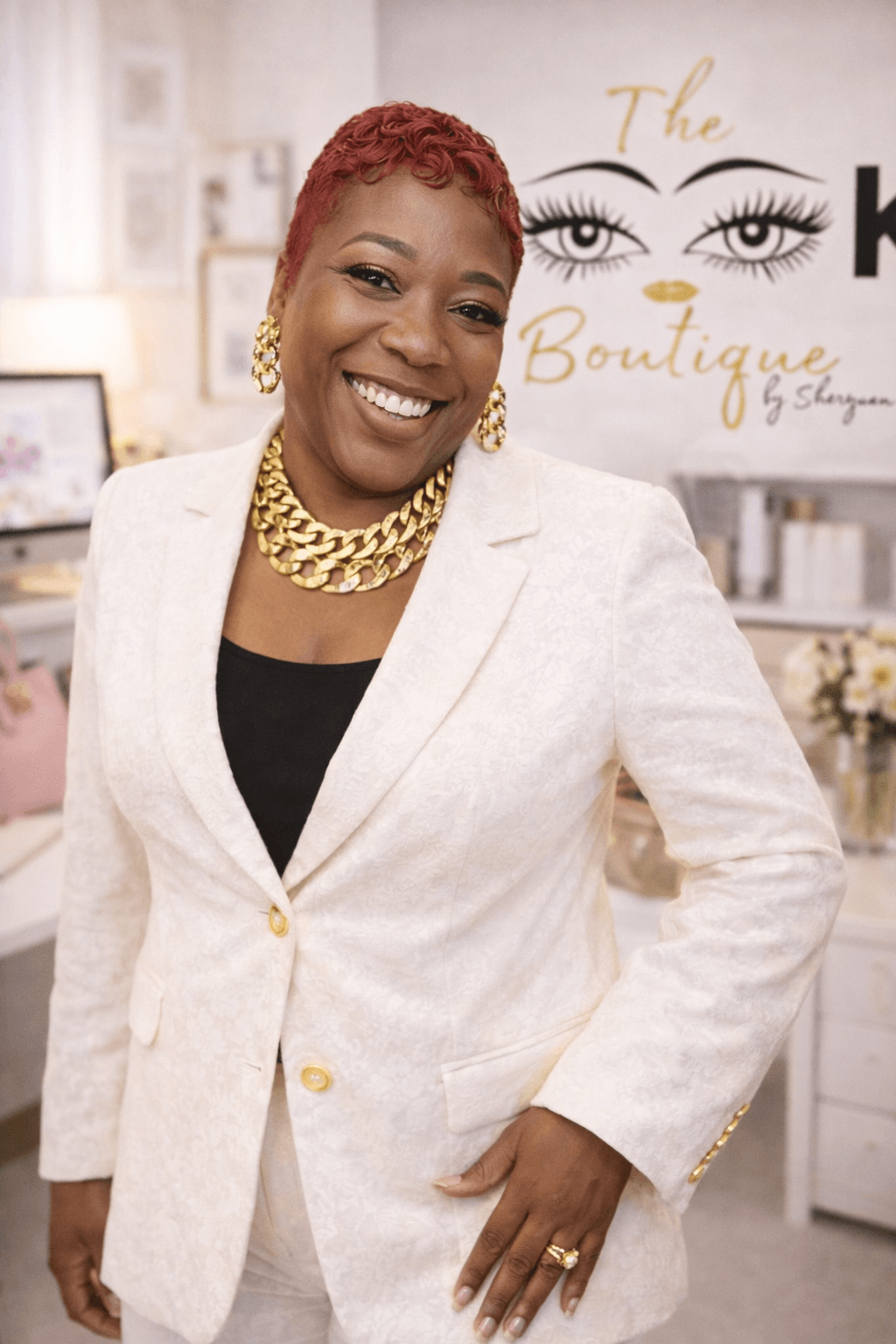 Woman with short red hair wearing a white blazer, gold jewelry, smiling in a boutique setting.